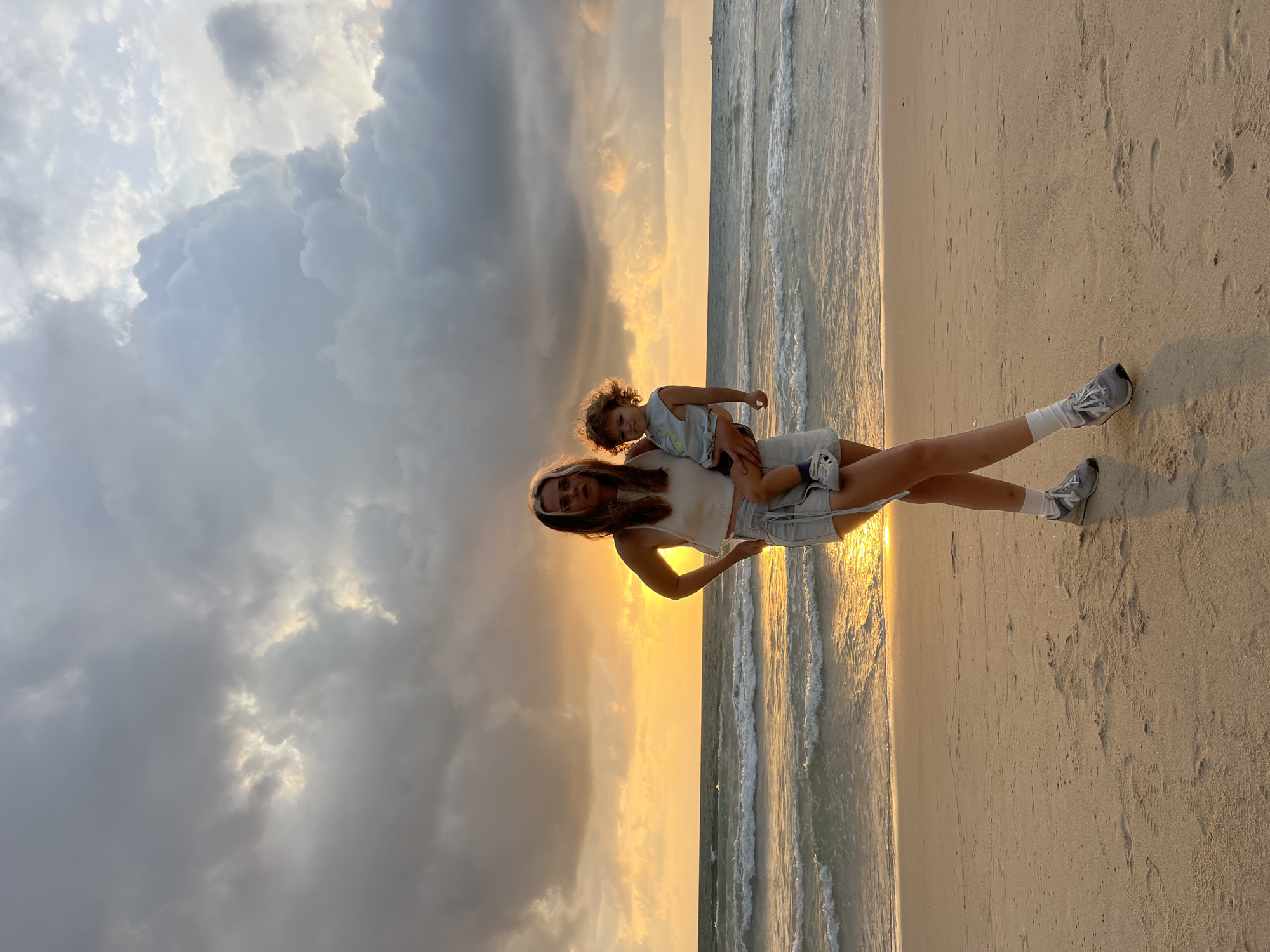 In the city of Choeng Thale, Thailand, a young woman stands on a sandy beach, holding a toddler. The woman is wearing a white tank top and light-colored shorts. Her hair is light brown with a streak of white. The toddler is dressed in a light blue tank top and is held in her arms. The ocean is to their left, with gentle waves rolling onto the shore. The sky is dramatic, filled with dark, heavy clouds illuminated by the golden light of a sunset. The sun casts a warm glow on the water and the sand. The overall scene is a moment captured during a sunset on the beach.