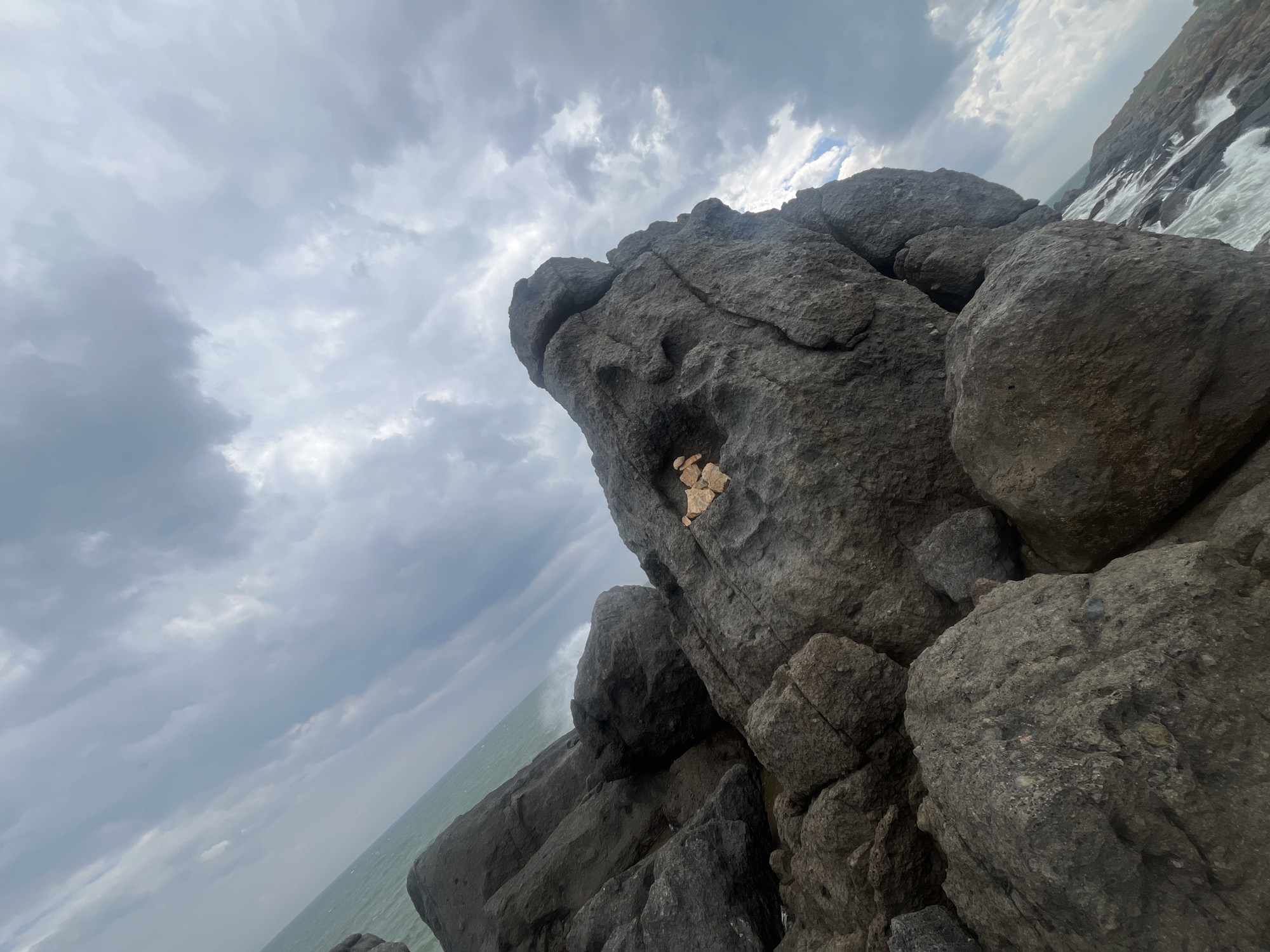 A low-angle, wide shot shows large, dark grey rocks along the coast of Mui Ne, Vietnam. The rocks are wet and textured, with some smaller, lighter-colored rocks stacked on a larger rock face in the center of the image. The ocean, with green water and white waves, is visible to the left, and a cloudy, grey sky fills the upper portion of the frame. The overall scene is dramatic and rugged, with the tilted perspective adding to the sense of scale.