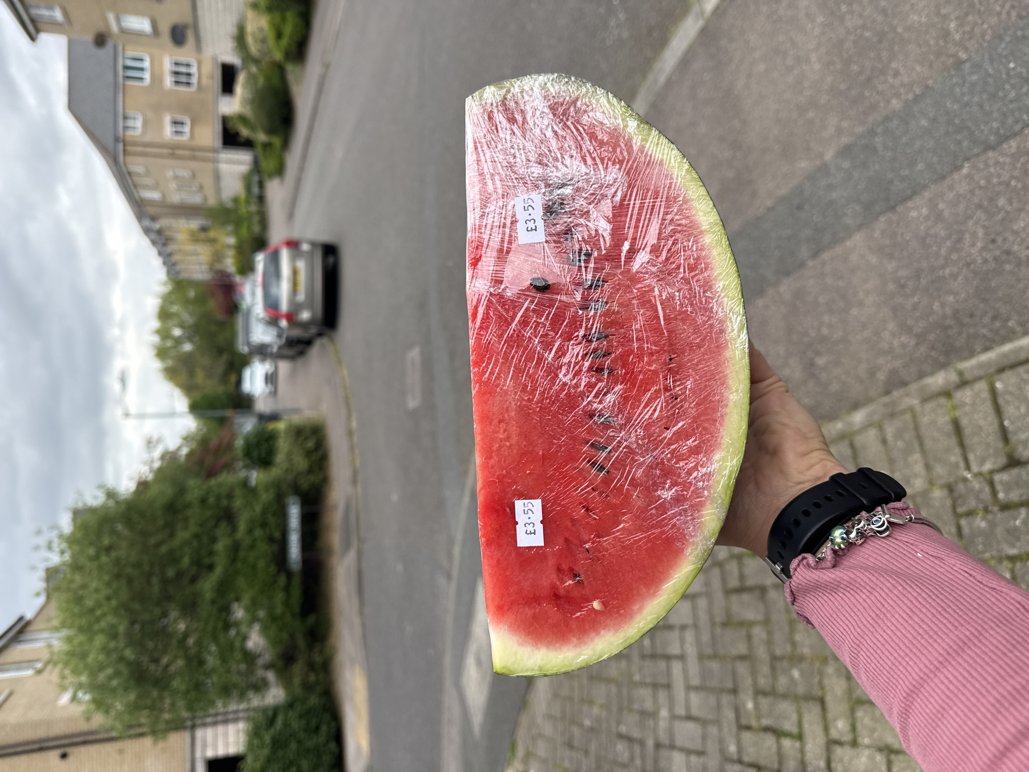 A medium shot shows a person holding a wrapped half watermelon outdoors, with a residential street scene in the background. The watermelon is wrapped in clear plastic and has two price tags reading "£3.55" attached to it. The watermelon flesh is red with black seeds and a green rind. The person holding the watermelon has a visible wrist with a black watch and silver bracelets. They are wearing a pink long-sleeved shirt. The background includes a blurred street with houses, trees, and parked cars. A street sign is partially visible. The street scene is consistent with a location in Great Yarmouth, United Kingdom.