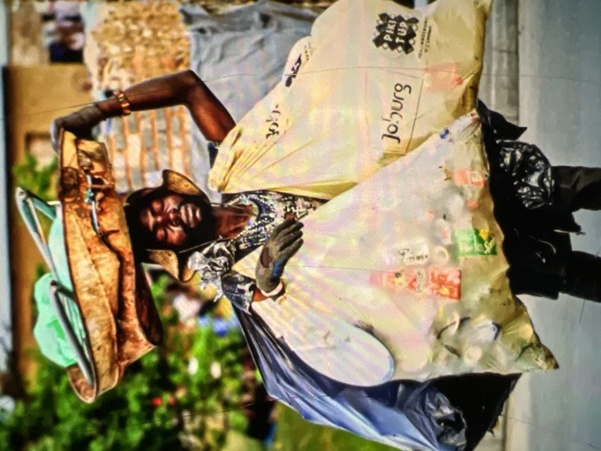 The image depicts a person walking through a city, carrying a basket on their head and large bags of what appears to be recyclables. The person is wearing a hat, gloves, and a necklace. The bags are transparent, revealing various items within, including plastic bottles and other discarded materials. One of the bags has the word "Joburg" printed on it. The background shows buildings and greenery, suggesting an urban environment. The presence of the word "Joburg" on the bag indicates the image may have been taken in Johannesburg, South Africa, rather than Lagos, Nigeria. The scene appears to be a snapshot of daily life, potentially showing a scavenger or waste collector at work.
