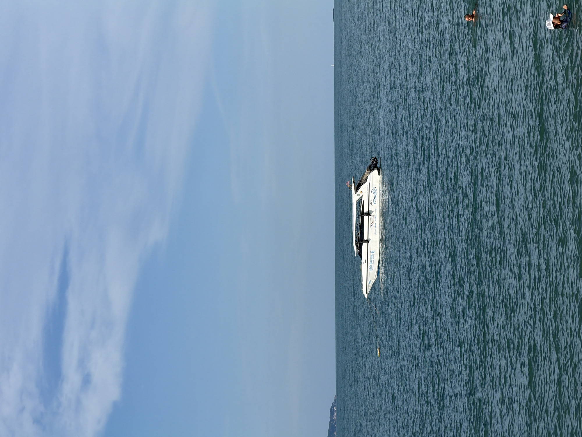 A white speed boat with a black roof is positioned on a calm, blue ocean under a clear sky with sparse white clouds. The boat is slightly to the left of the center of the frame, and its wake is visible. The boat has writing on its side in blue, and a flag is flying from its rear. A yellow marker buoy is floating to the left of the boat. Two people are visible in the water to the right of the boat, one partially submerged and the other with their head and shoulders above the water. In the far distance, a faint outline of land is visible on the left horizon, and a distant ship is present on the right horizon. The scene appears to be a coastal area in Baan Tai, Thailand.