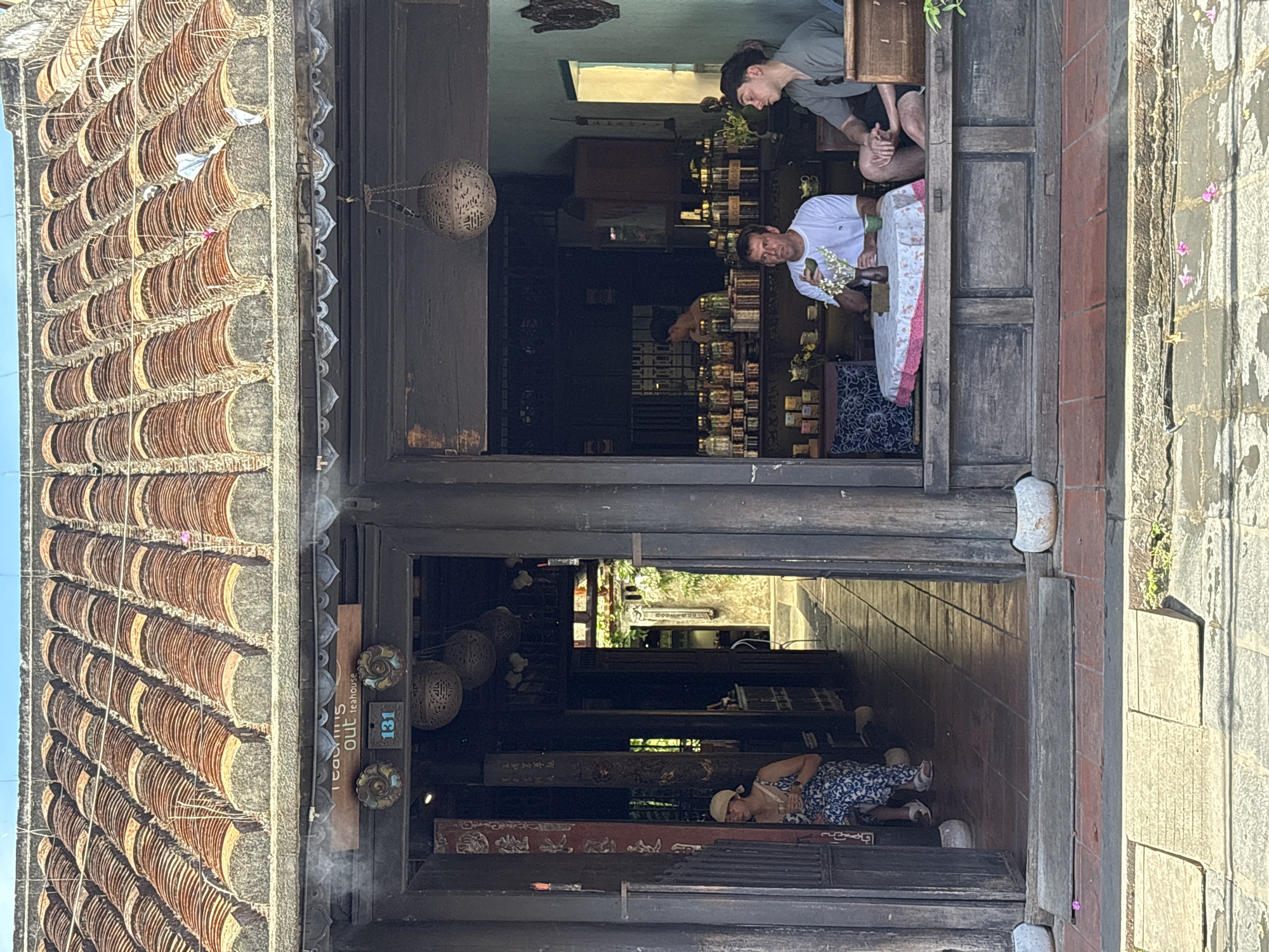 This image depicts the entrance to a teahouse in Hội An Ward, Vietnam. The teahouse has a traditional tiled roof and wooden facade. Two men are seated at a table inside the teahouse, one holding a teacup and the other looking out. A woman is seated to the left of the entrance. Shelves stocked with jars of tea are visible in the background. The street in front of the teahouse is paved with stone.