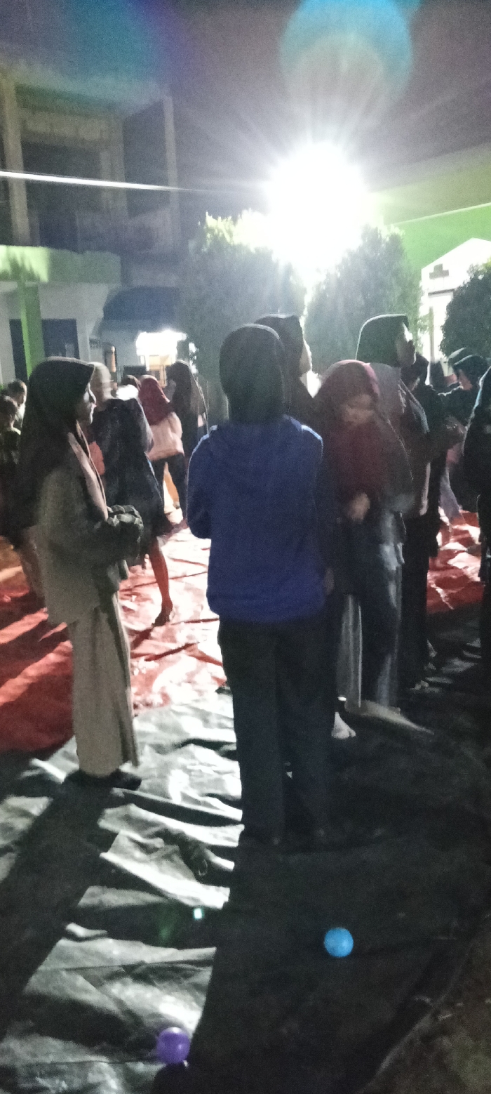 The image shows a group of people standing on a black tarp in front of a building at night. The people are standing in a dimly lit area with a large light source shining brightly in the background. The light source creates a glare that obscures some of the details of the people and the surrounding environment. There are eight people in the image. The people are dressed in dark clothing with the exception of one person wearing a light blue jacket and another wearing a red scarf. The people appear to be talking and interacting with each other. There are two small, colorful balls on the ground in the foreground of the image, one blue and one purple. The image was taken in Cangkuang, Indonesia, at coordinates -7.065 latitude, 107.552 longitude. The image is taken from a low angle, looking up at the people. The background is blurry due to the light source. The image is slightly out of focus, but the overall scene is clear.