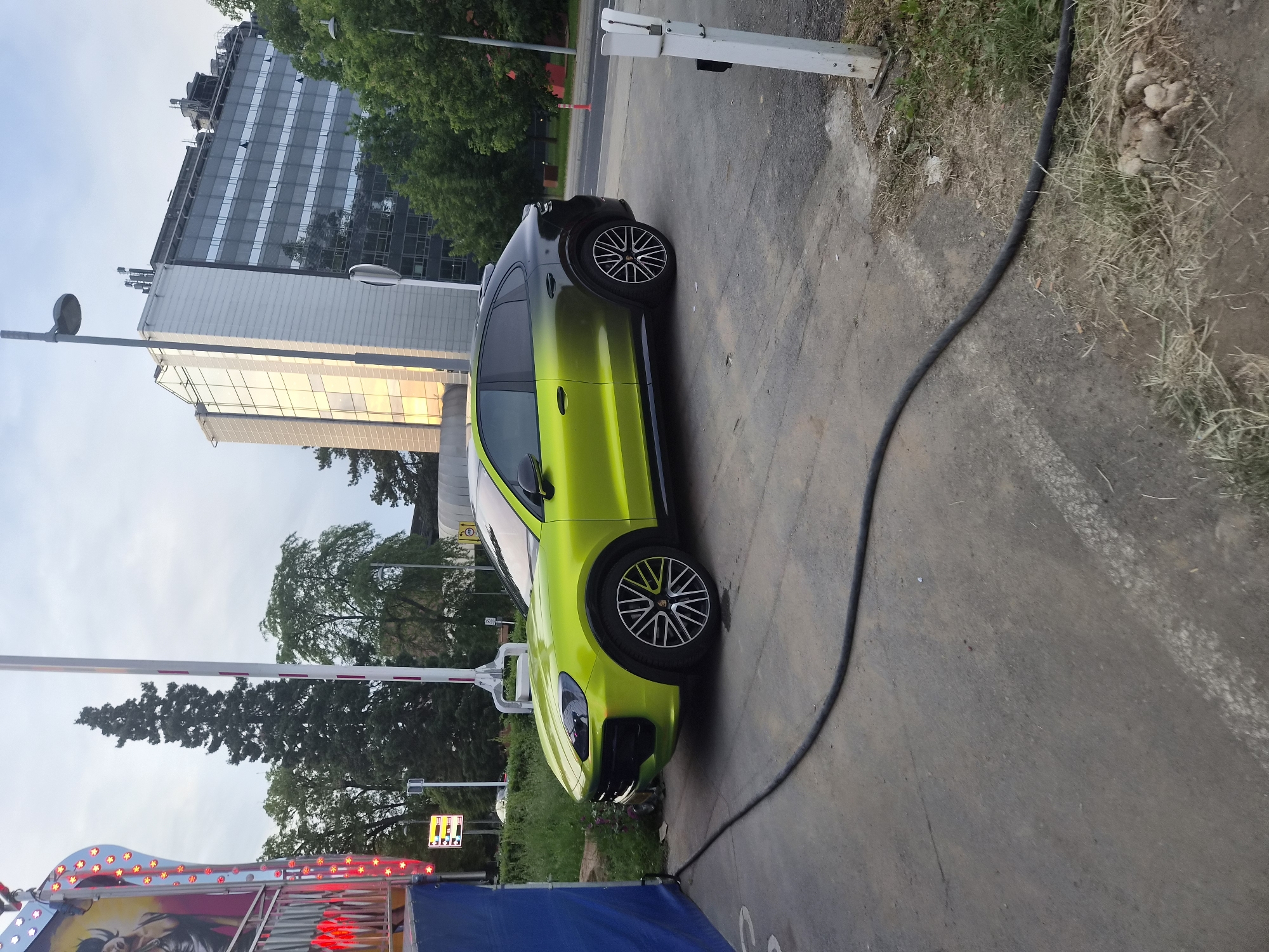 The image captures a green Porsche Cayenne parked on the side of a road in Esch-sur-Alzette, Luxembourg. A large modern building and trees are visible in the background. A black cable runs along the edge of the pavement near the car. In the foreground, a portion of a fairground ride with blue and red lights is visible.