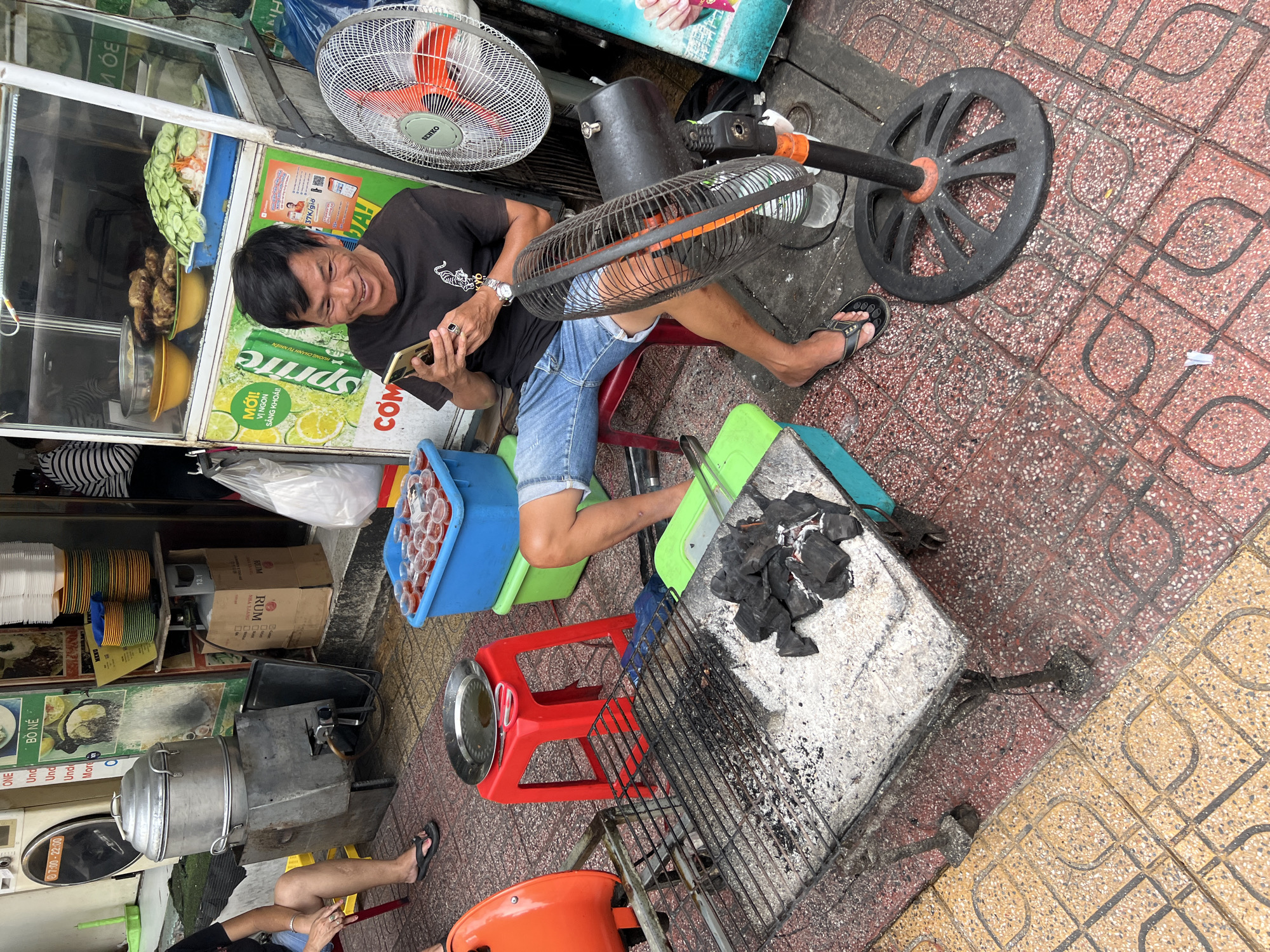 The image depicts a street food vendor in Nha Trang, Vietnam, actively engaged with his mobile phone while seated at a makeshift stall. In the foreground, a charcoal grill is visible, covered in ashes and scattered pieces of charcoal. Adjacent to it, a bright orange plastic chair is positioned, with a red stool and a metal tray on top. Behind the grill, a woman's legs are visible, suggesting she is a customer or passerby. A large metal steaming apparatus, likely used for cooking, is present to the left, with a gas cylinder attached. The vendor himself, a middle-aged man with short dark hair, is seated on a red plastic stool, wearing a black t-shirt and denim shorts, engrossed in his smartphone. He is smiling, indicating a relaxed demeanor.
The background features a food stall with a glass display case showcasing various food items, including what appears to be fried chicken and a large platter of sliced cucumbers. Colorful stacks of plates are arranged on shelves, and a green Sprite can is prominently displayed. A large white fan stands to the right of the vendor, its blades partially obscured by a smaller black fan. The ground is paved with textured tiles in shades of red and yellow. The overall scene suggests a typical bustling street scene in Nha Trang, characterized by outdoor food vending and casual interactions.