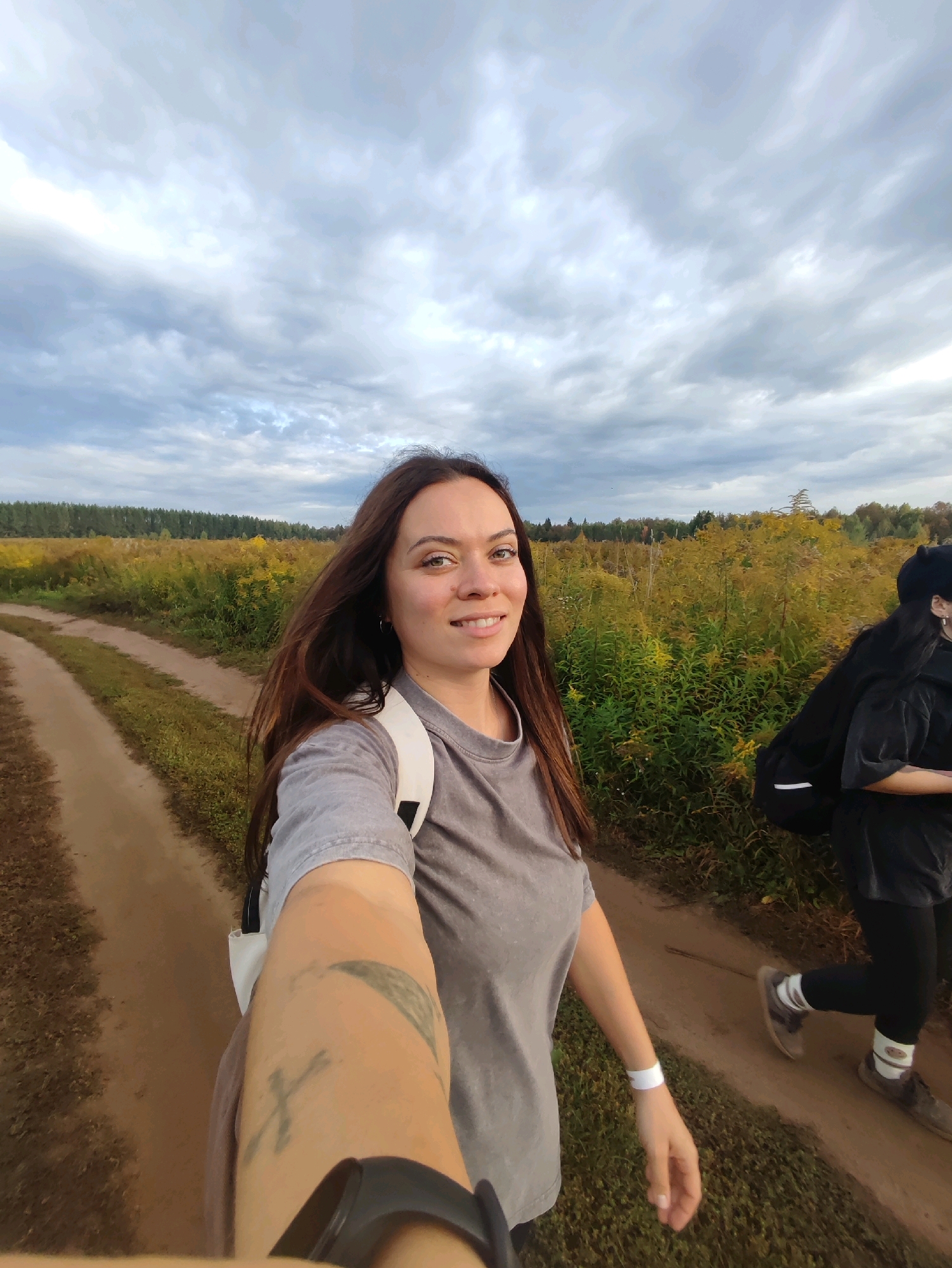 An outdoor selfie is taken from a low angle, capturing a young woman from the chest up as she smiles at the camera. She has long, dark brown hair and is wearing a grey t-shirt and a white backpack. Her left arm is extended forward, holding the camera, and a tattoo is visible on her forearm. A white wristband is also on her left wrist. To her right, another person is partially visible from the side, wearing black clothing and a black hat. They appear to be walking away from the camera on a dirt path. The path runs through a field of tall, yellow and green vegetation, likely wildflowers, with a line of trees in the distance. The sky above is filled with dramatic, cloudy formations. The scene is set in Vozhoiskii, Russia.