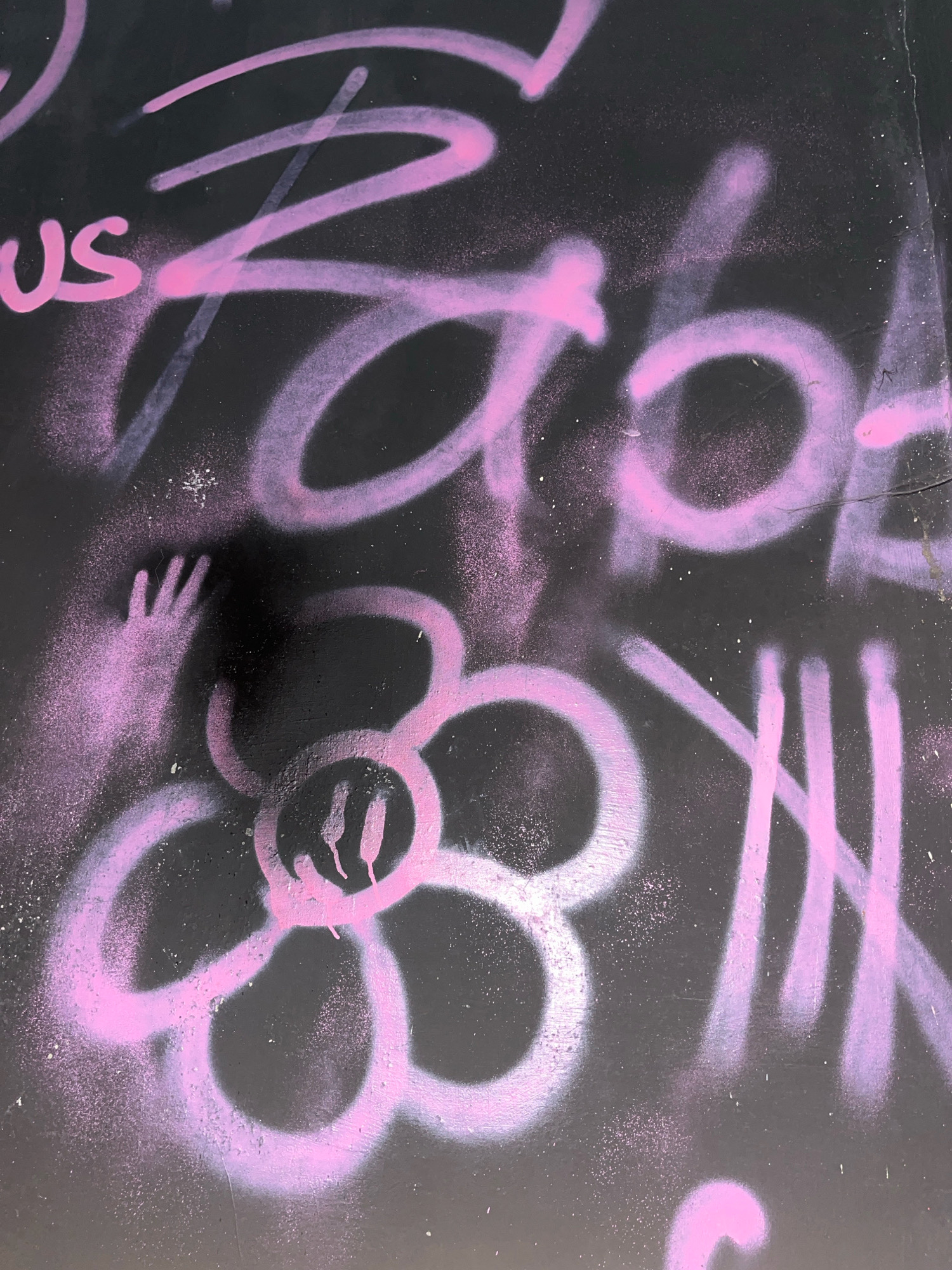 The image shows graffiti spray-painted in pink on a black surface, possibly a wall or a metal panel. The graffiti includes text, a handprint, and a flower with a smiley face. The text is illegible due to the style and partial visibility. The handprint is a silhouette of an open hand with spread fingers, appearing to be reaching out. The flower has five petals and a simple face with two vertical lines for eyes and a curved line for a smile. The overall style of the graffiti is consistent with street art. This graffiti is located on a wall in the city of Surakarta, Indonesia.