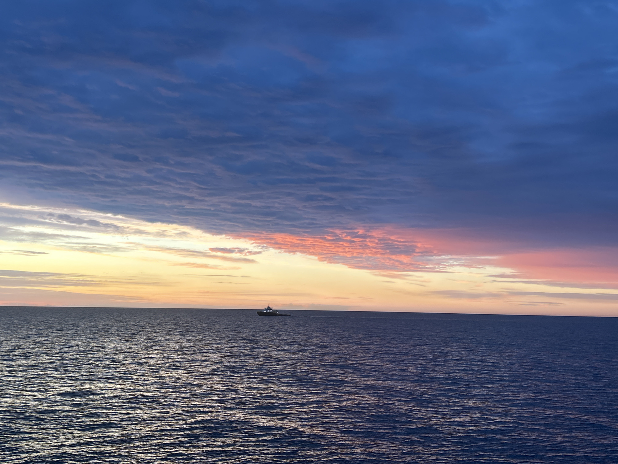 A wide shot captures a serene ocean scene at dusk. The sky is a gradient of deep blues and purples, transitioning to streaks of pink and orange near the horizon. Wispy, textured clouds fill the upper half of the frame. The sea below is a dark, choppy expanse of deep blue, reflecting the colors of the sky. In the middle distance, a small, solitary vessel, possibly a tugboat or workboat, sails across the water. The boat is silhouetted against the lighter sky. The horizon line is sharp and distinct, separating the sea from the sky. The image conveys a sense of calm and solitude.