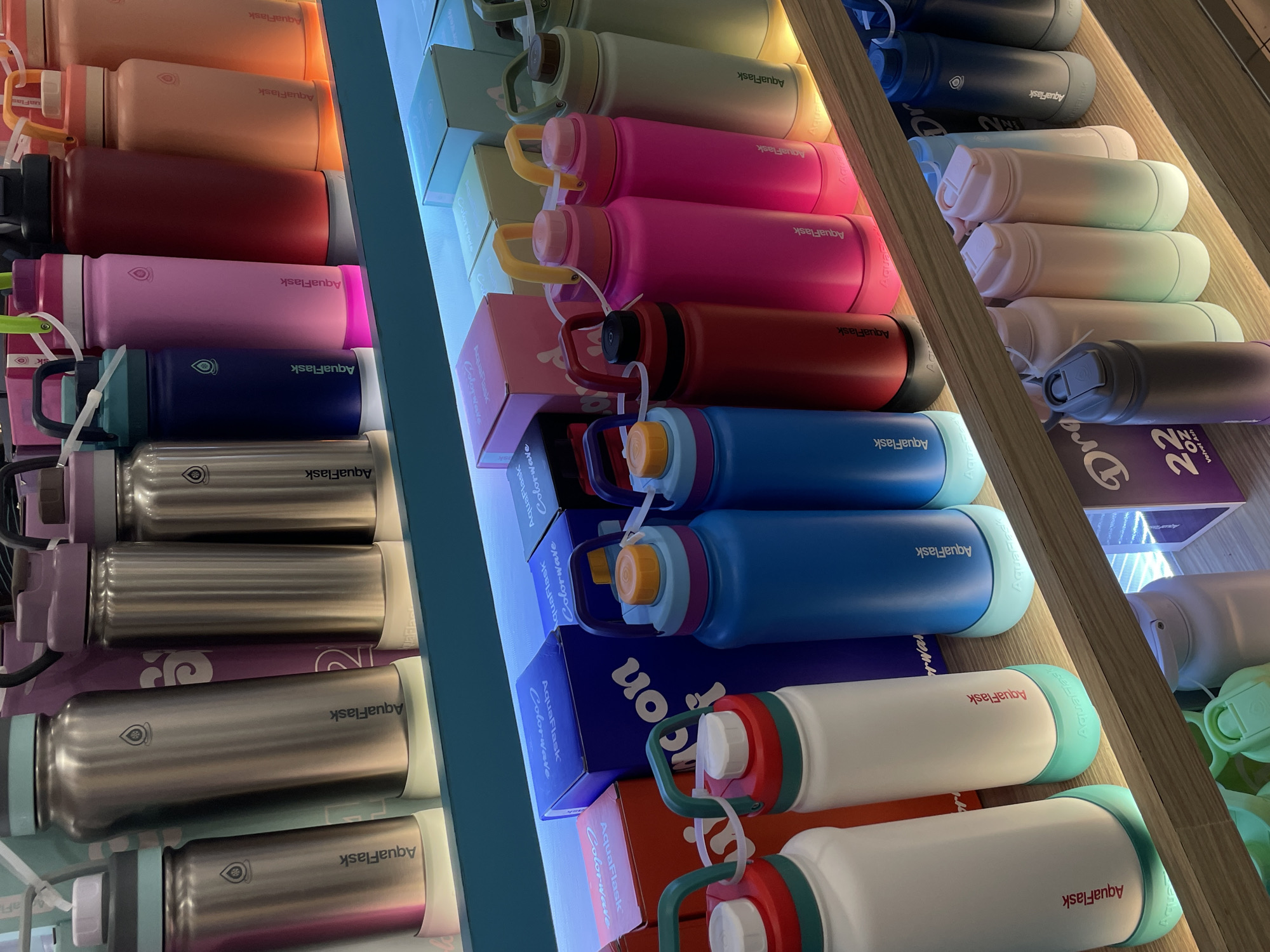 This image depicts a display of AquaFlask brand water bottles on shelves in a retail setting, likely a store. The bottles are of various colors including silver, blue, pink, red, peach, cream, and light green, and many feature the AquaFlask logo. Some bottles are standing upright, while others are partially obscured. Boxes with "AquaFlask Colorwave" branding are also visible behind some of the bottles. The lighting is bright, highlighting the products. The scene is clean and organized, typical of a retail environment. There are no people visible in the image.
Without further information, it is impossible to definitively pinpoint the location as Davao City, Philippines. However, if this image were taken in Davao City, it could be from a store within a shopping mall such as SM Lanang Premier, Abreeza Mall, or Gaisano Mall, or a standalone AquaFlask branch. Such retail displays are common in these types of establishments in Davao City.