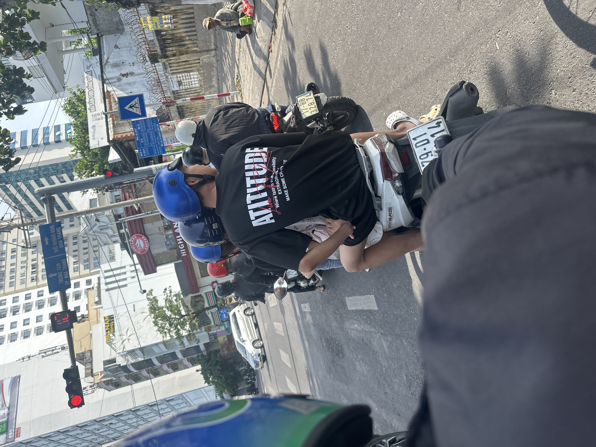 The image captures a busy street scene in Bac Nha Trang Ward, Vietnam, likely during the daytime given the strong sunlight and shadows. The perspective is from someone riding on the back of a motorcycle, looking forward at other people on motorcycles and a white car.
The primary subjects are several individuals on motorcycles, wearing helmets of various colors, including blue, white, and red. One person in the foreground is wearing a black t-shirt with the word "ATTITUDE" prominently displayed in white lettering, accompanied by smaller text. Another individual is wearing a black jacket and jeans. The motorcycles themselves are varied, with one featuring a license plate that reads "79-01 14".
The setting is an urban environment with buildings lining the street. One building has a sign that says "DAL NAM HOTEL" and another advertises "HIGHLAND COFFEE". A traffic light is visible, showing a red signal. There are also street signs, including one with Vietnamese text that appears to indicate directions to "TRUNG TÂM TP. NHA TRANG" (Center of Nha Trang City) and "SUỐI KHOÁNG NÓNG NHA TRANG" (Hot Mineral Springs Nha Trang). Another sign reads "HẢI SẢN TƯƠI SỐNG GIÁ RẺ" (Fresh Seafood Cheap Price).
The activity appears to be typical urban traffic. The overall mood is functional and everyday, with no strong emotional cues evident from the distant figures. The presence of multiple vehicles and pedestrians suggests a moderately populated area. The weather appears to be sunny and clear.