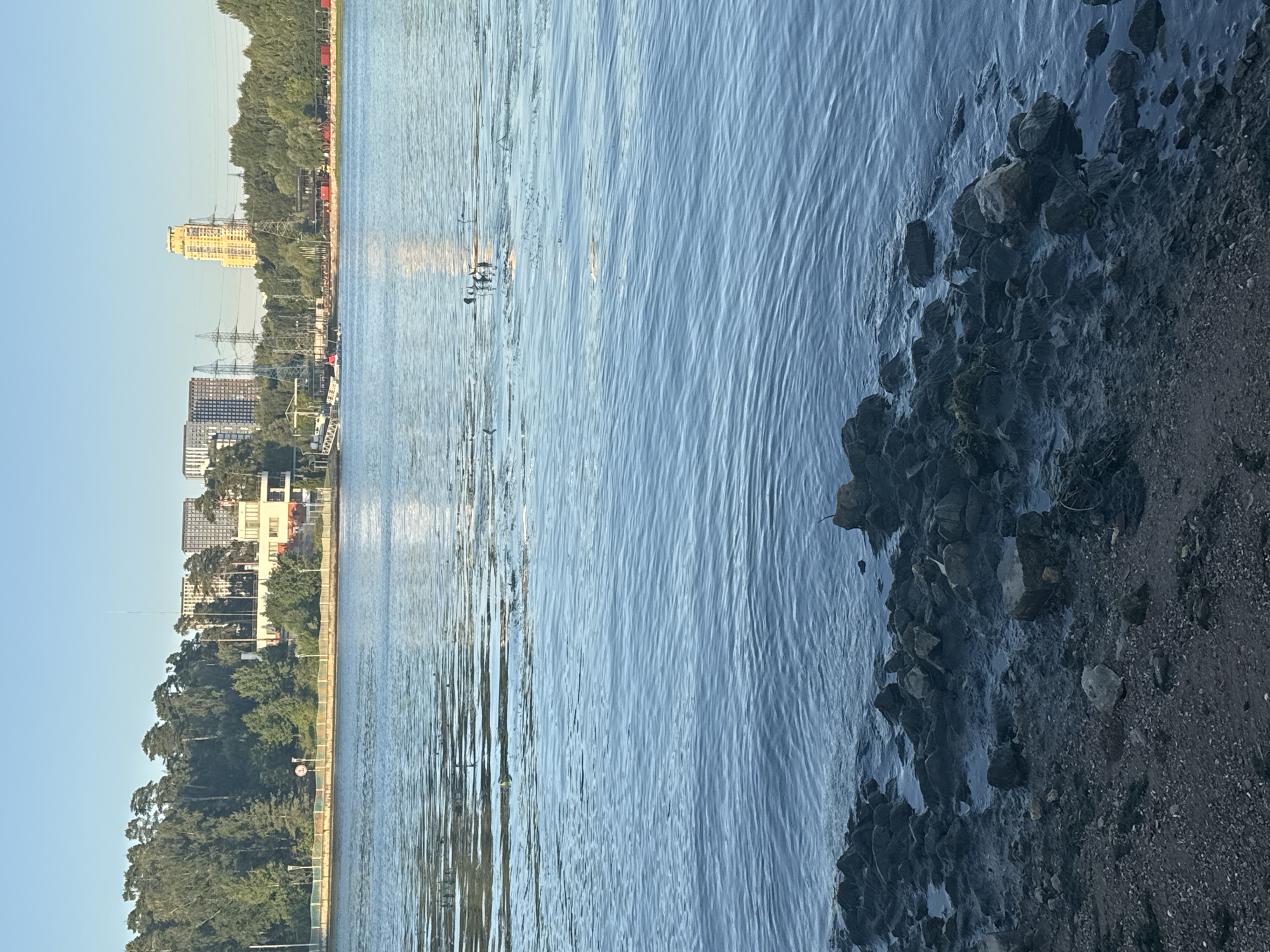The image depicts a wide, calm body of water, likely a river or reservoir, with a rocky, somewhat muddy shoreline in the foreground. The water reflects the clear blue sky and the sunlight. In the mid-ground, a line of trees and vegetation borders the opposite bank. Beyond the trees, a cityscape is visible, characterized by modern buildings of various heights and architectural styles, including residential towers and other structures. Power lines and electrical pylons are also present in the distance, indicating an urban or semi-urban environment. The overall scene suggests a serene natural setting within or near a populated area. Based on the presence of the Klyazma Reservoir and the Moscow Oblast, this scene is consistent with the waterfront areas of Khimki, Russia, potentially near the Moscow Canal or a similar waterway.