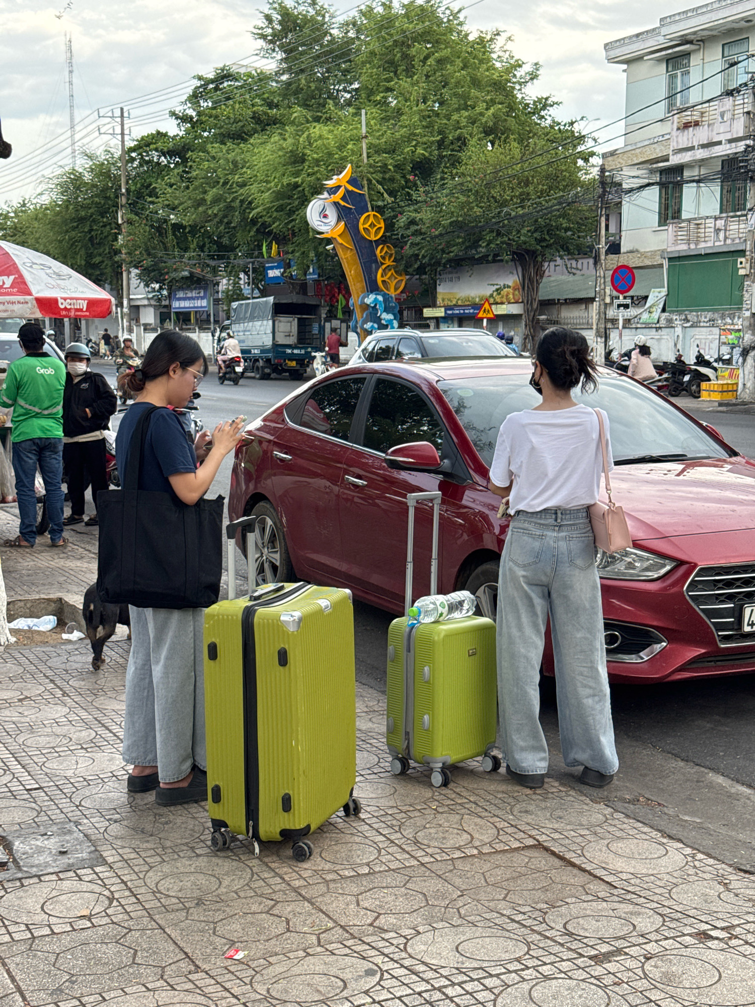 In Nha Trang, Vietnam, on a cloudy day, two young women stand beside a red sedan on a sidewalk. The woman on the left wears a dark blue t-shirt, grey pants, and carries a large black tote bag. She holds a phone in her hands and has a bright green suitcase at her feet. The woman on the right wears a white t-shirt, light blue wide-leg jeans, and a pink crossbody bag. She also has a bright green suitcase next to her, with a bottle of water on top. A black dog stands near the woman on the left, facing away from the camera. In the background, a street scene unfolds with traffic, trees, buildings, and a prominent telecommunication tower. Several other individuals and vehicles are visible, including a person in a green Grab uniform and a truck. The sidewalk is paved with patterned tiles.