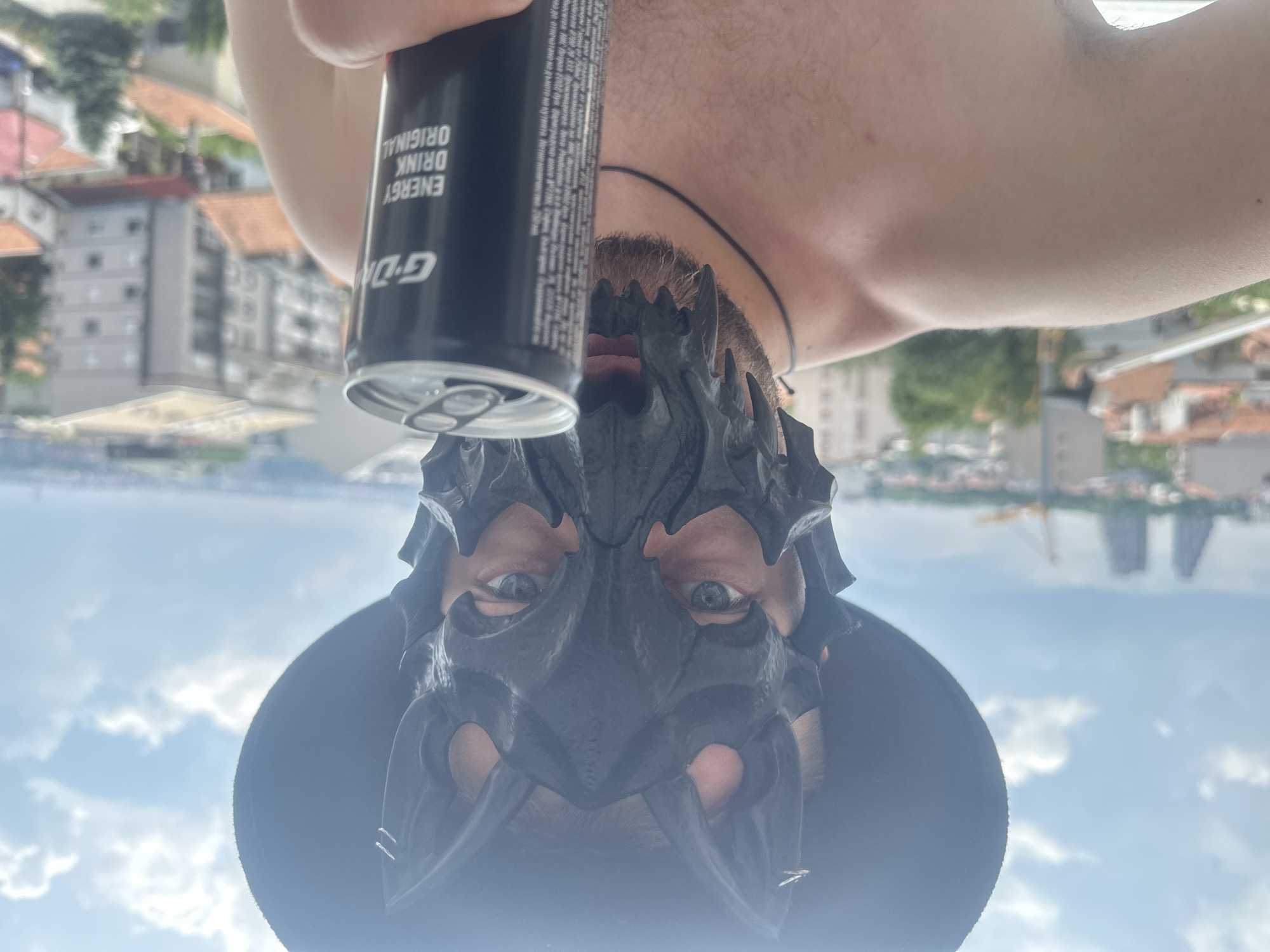 The image, taken outdoors, is upside down and features a person wearing a black mask and a hat. The person is holding a can of "G-D ENERGY DRINK ORIGINAL". The background shows buildings and a body of water under a cloudy sky, suggesting an urban or suburban setting with a water view. The lighting indicates it is daytime.