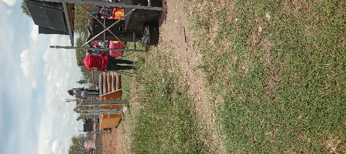 The image shows a scene in Kitengela, Kenya, at the coordinates -1.471 latitude and 36.958 longitude. There are three adults in the foreground, two women and one man. The women are wearing casual clothes and are standing on a set of stairs. The man is wearing a red shirt and is standing on the ground near the stairs. He is looking up at the women. There are a few other people in the background, sitting and standing. They are all casually dressed. The scene appears to be a gathering or event with a stage in the background. It is set up with a large, black speaker system placed on a wooden platform supported by a metal frame. There is a large, orange structure next to the stairs, and there is a metal structure used to support the speakers. The ground is covered in green grass with some bare patches and dirt. There is also a black bag on the ground near the speaker system. There is a large, open grassy area in the foreground with a cloudy sky. The image was likely taken during the day.