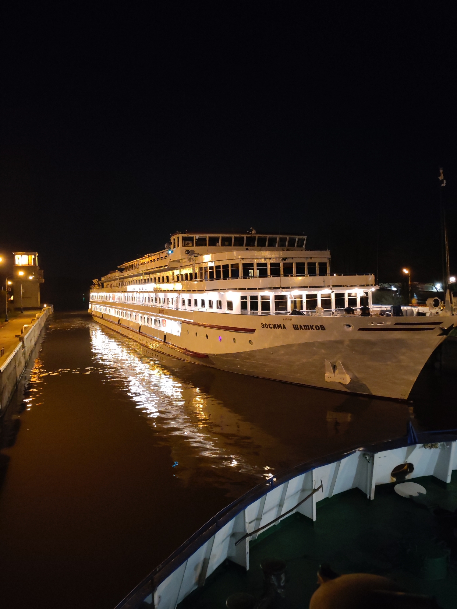 At night, the passenger ship "Zosima Shashkov" is traveling through a canal in Yakhroma, Russia. The ship's lights are illuminated. The canal's water reflects the ship's lights. The ship is headed straight. The ship's name, "ZOCUMA WAWKOB" is printed on its side. Part of a nearby ship is visible in the foreground. Buildings are visible in the background.