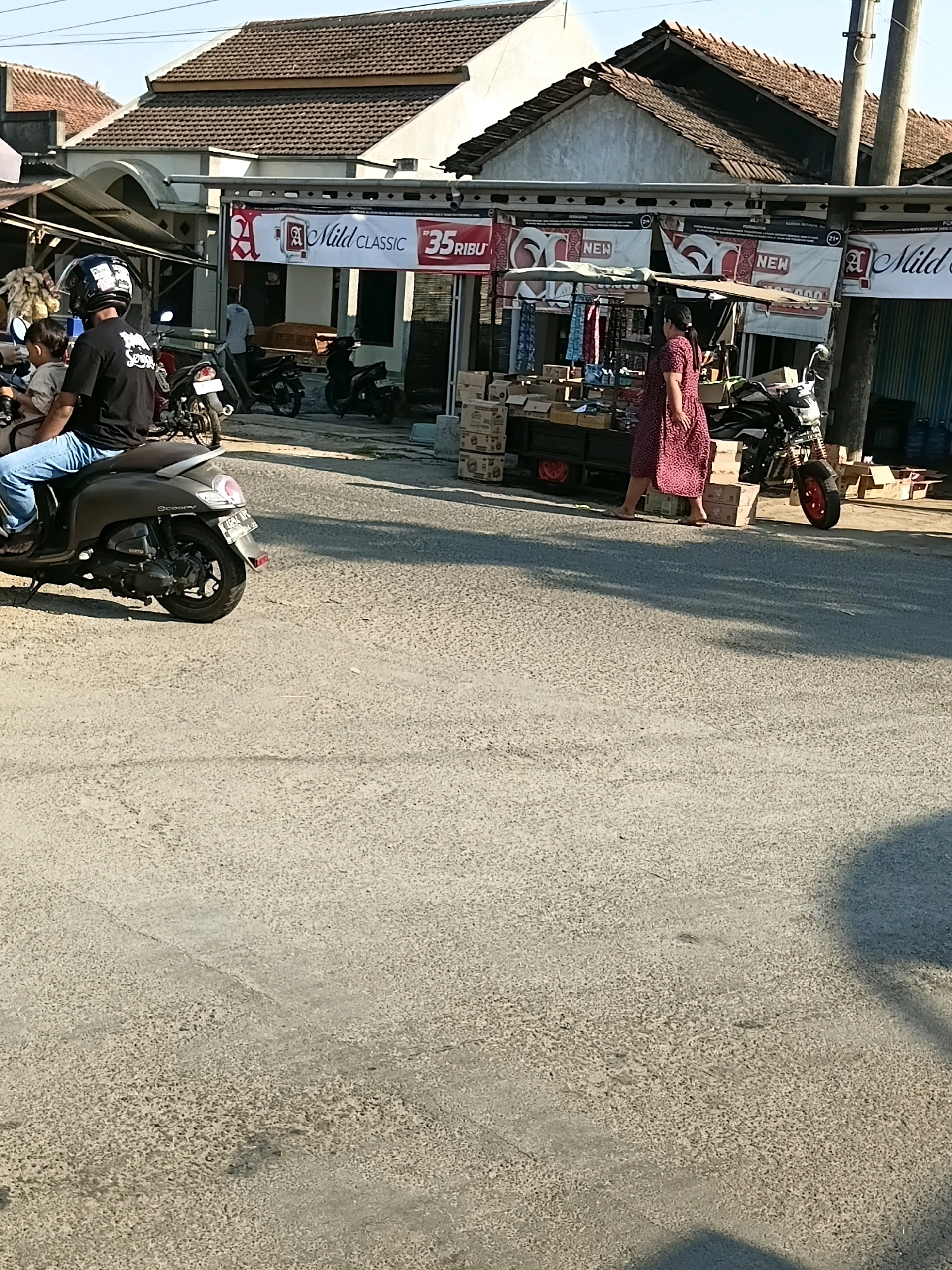 In Cikal, Indonesia, a busy street scene unfolds under a bright sky. In the foreground, a person rides a dark-colored scooter with a child on the back, both wearing helmets. The scooter has a license plate that reads "L 5354 W". Beside the road, a woman in a patterned dress walks past a market stall laden with goods and cardboard boxes. Behind the stall, a motorcycle with red wheels is parked. The market is situated in front of a building with a tiled roof and banners displaying "A Mild CLASSIC" and "35 RIBU". Further down the street, other motorcycles are parked, and people are milling about. The road surface is asphalt, textured and showing signs of wear. The buildings in the background are simple structures with tiled roofs, typical of Indonesian architecture. The overall impression is of a vibrant, everyday scene in a lively Indonesian town.
