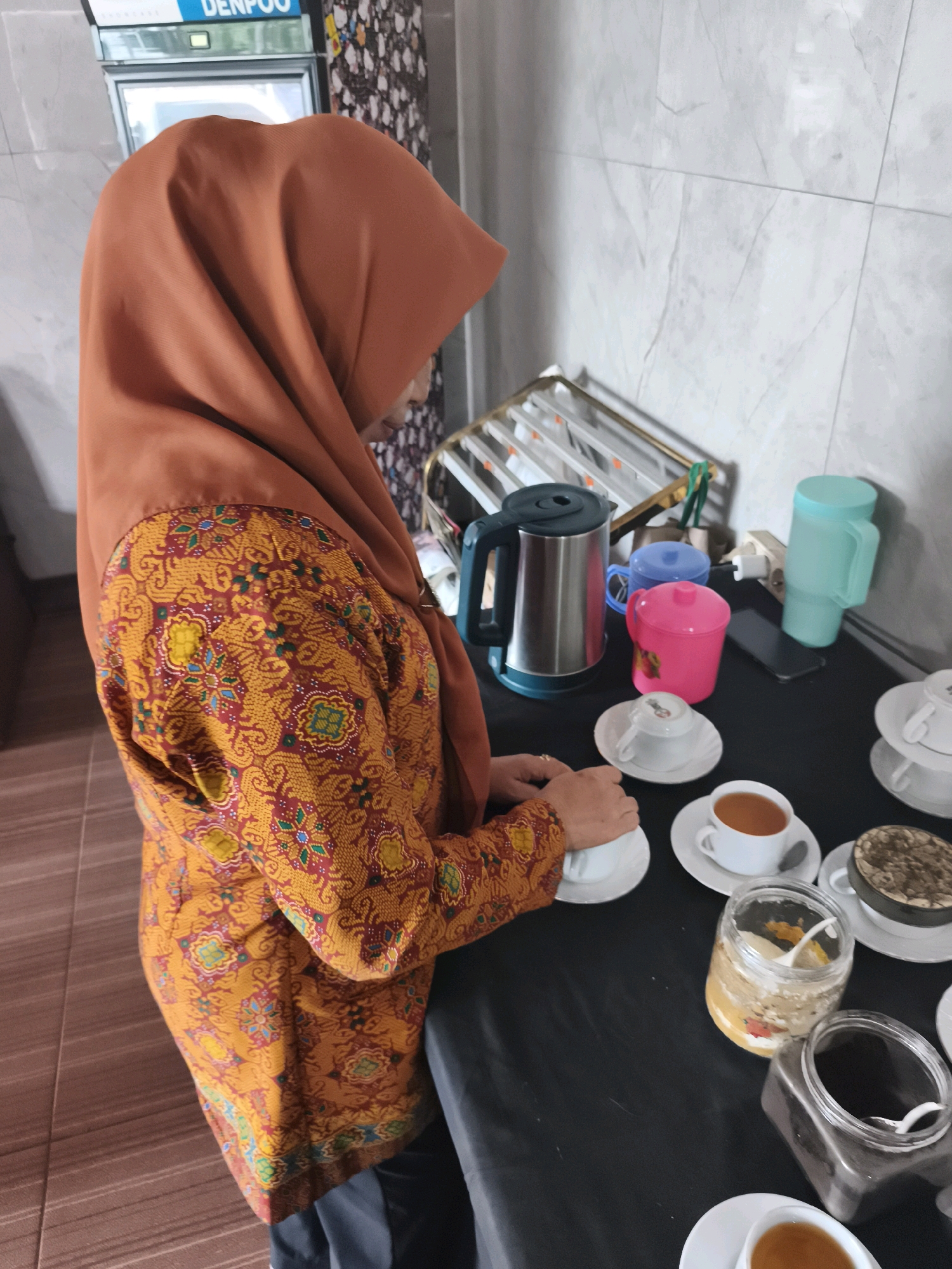A woman wearing a burnt orange hijab and an orange and red batik patterned long-sleeved top stands on the right side of the frame, preparing or serving tea. She is positioned in front of a black table laden with tea service items. The table displays multiple white teacups and saucers, some containing amber-colored liquid, likely tea. A silver electric kettle with a teal base and lid sits on the left side of the table, accompanied by a pink lidded cup and a blue lidded cup. To the right of the kettle are two larger, colored plastic pitchers, one turquoise and one pale green. Two glass jars are also present on the table; one contains a light brown granular substance, possibly sugar or creamer, with a small spoon, and the other, a darker granular substance with a spoon. The background features a tiled wall with a marble-like pattern and a rack with several items. A refrigerator with "DENPOO" branding is visible in the upper left corner. The floor is tiled with a wood-grain pattern. The scene suggests a domestic or hospitality setting, possibly in Palembang, Indonesia, given the prevalence of batik attire and the "Denpoo" brand which is commonly found on refrigerators in Indonesia.