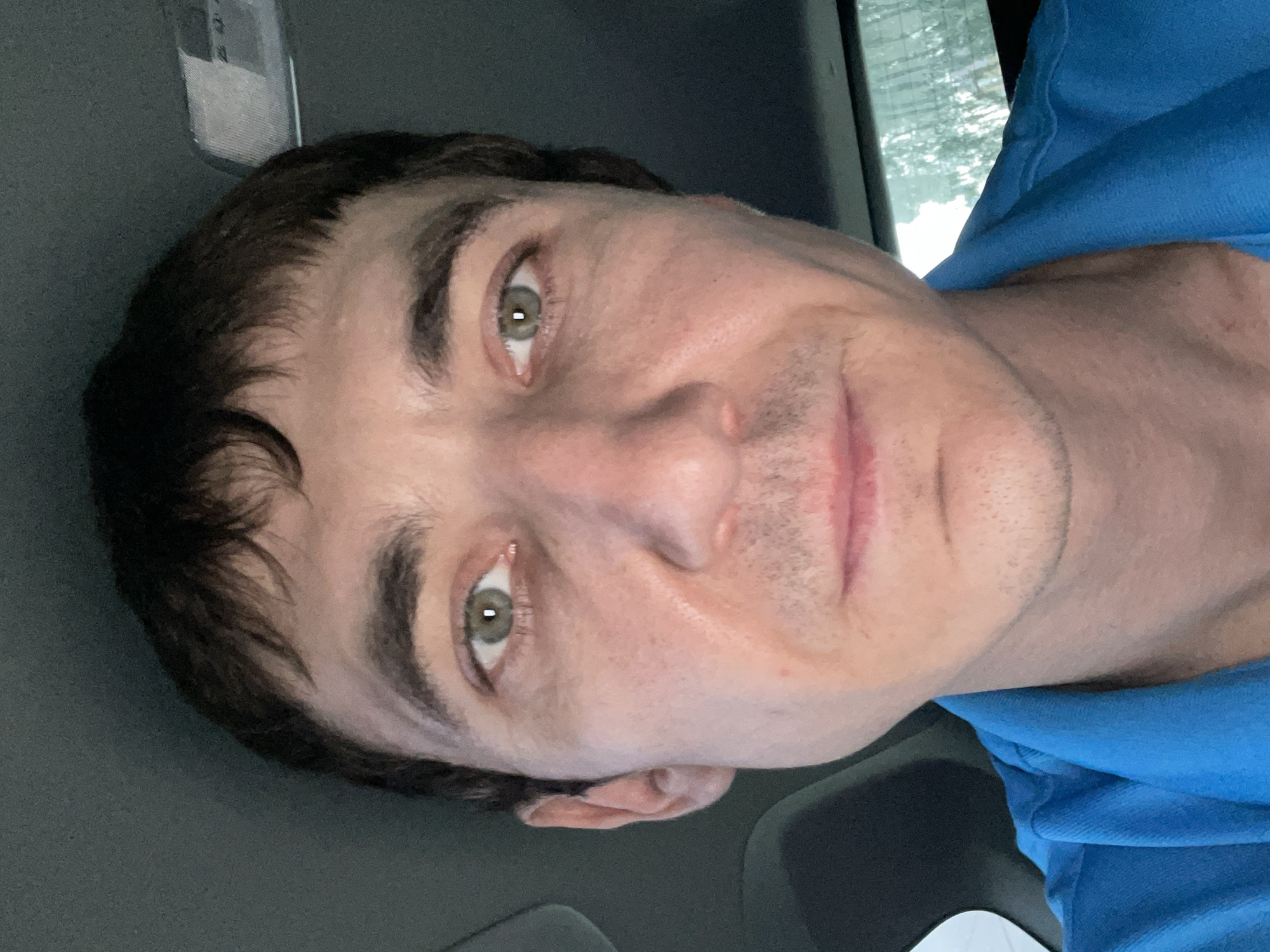 The image shows a close-up, selfie-style portrait of a man with fair skin, dark hair, and green eyes. He is wearing a blue collared shirt. The background appears to be the interior of a car, with hints of the car's ceiling and what looks like a rear window with trees visible outside. There is no indication of the city of Laishevo, Russia.