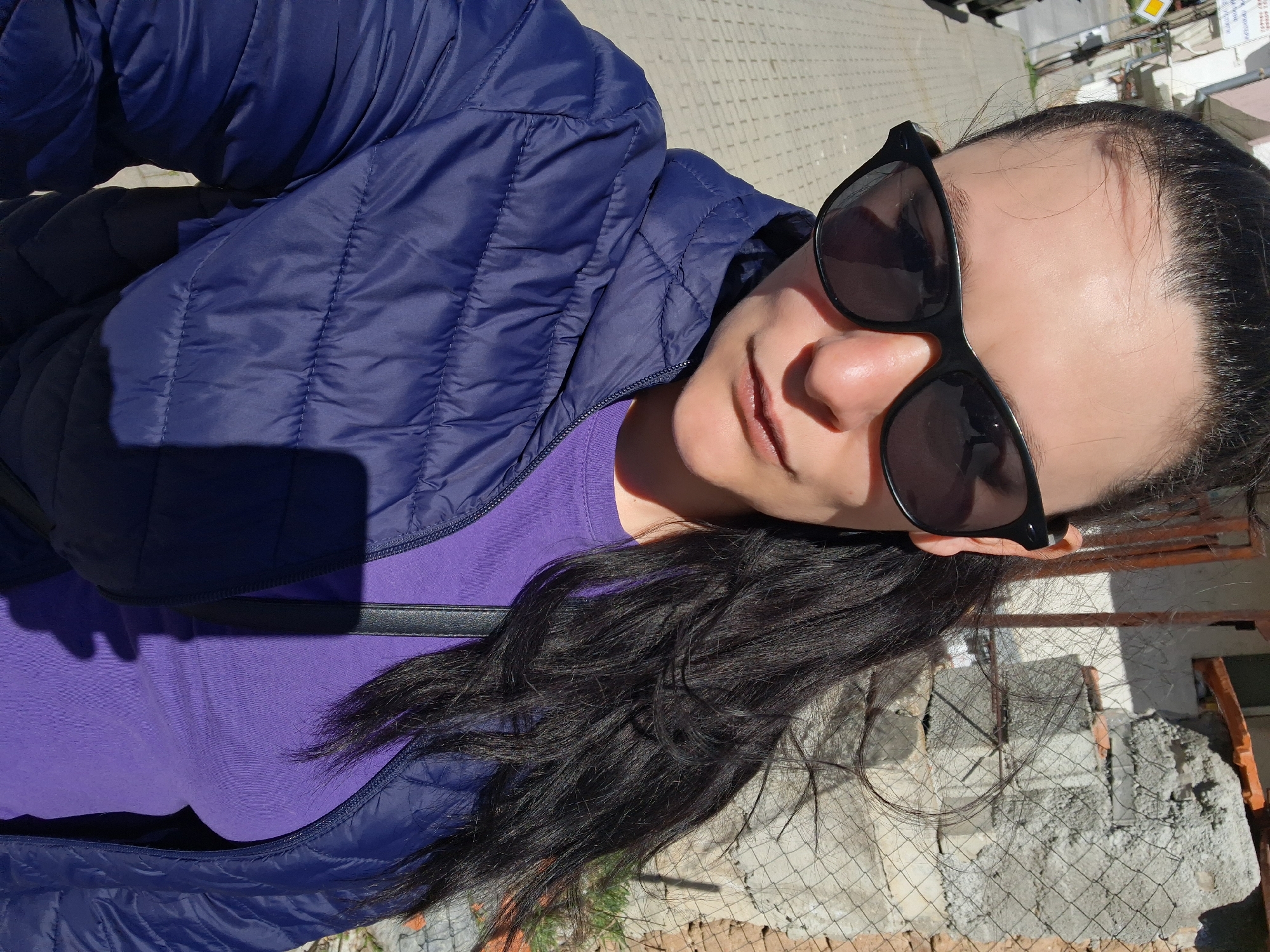 A woman is posing outdoors, likely in Goce Delcev, Bulgaria. She is wearing a blue quilted jacket over a purple t-shirt and sunglasses. Her dark hair is pulled back. The background includes a brick or concrete wall and some fencing, suggesting an urban or semi-urban environment.