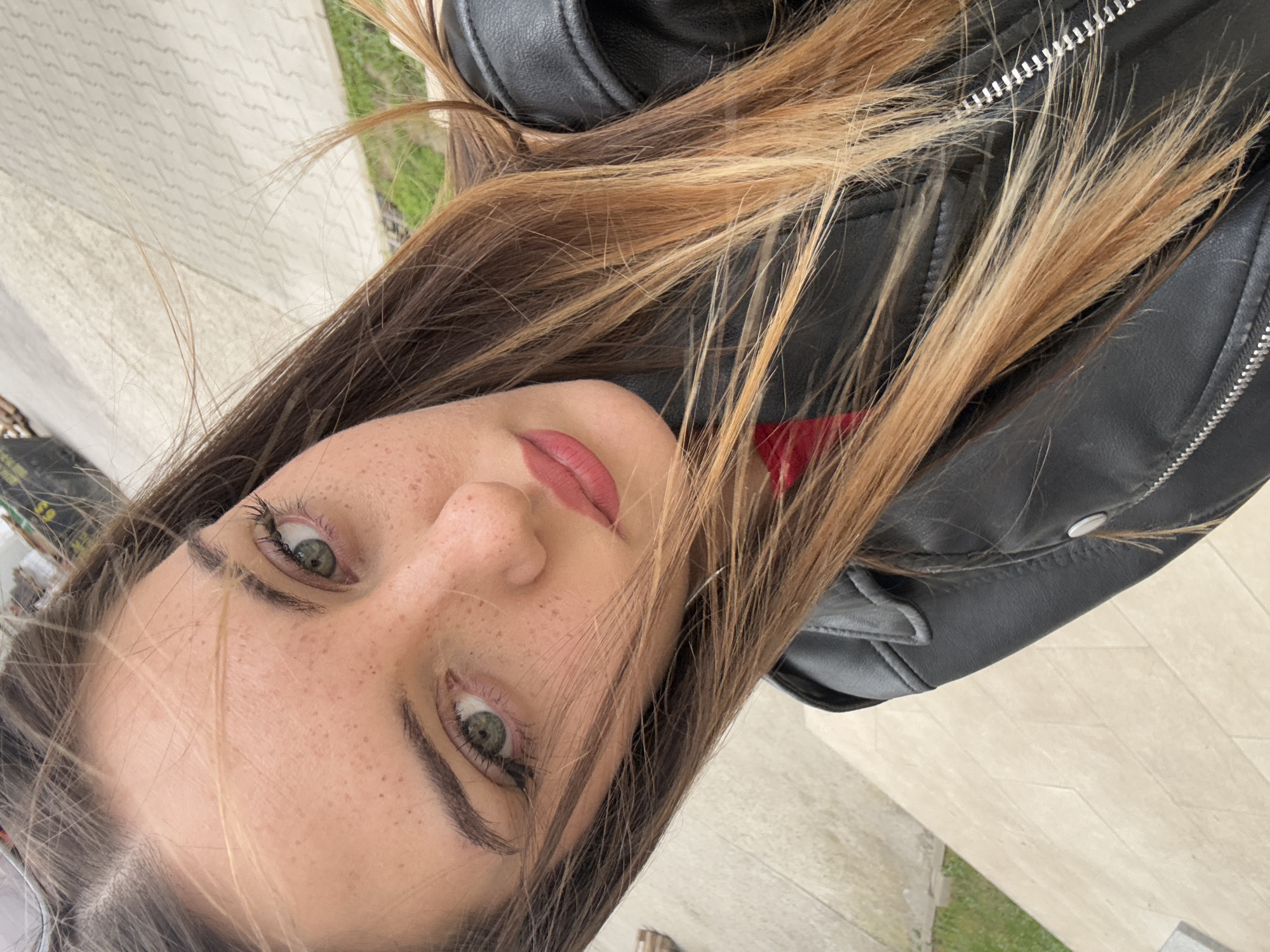 A close-up, angled selfie photograph captures a young woman with light skin, freckles, and green eyes. She has brown hair with blonde highlights. She's wearing a black leather jacket. The background includes a tiled surface and a hint of green vegetation. No specific landmarks are visible to definitively confirm the location within Sofia, Bulgaria. No actions or interactions are happening within the scope of the image, as it solely focuses on the subject's self-portrait.