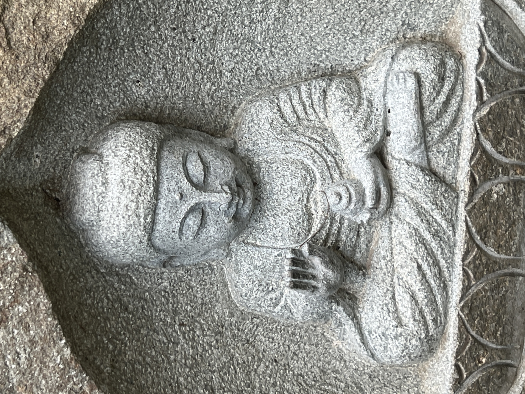 A close-up, eye-level, vertical shot shows a grey stone carving of a seated Buddha figure recessed within a rough, textured grey stone background. The Buddha is depicted in a meditative pose, with eyes closed, a calm expression, and hands resting in its lap, holding a small object. The carving details the Buddha's robe, hair, and facial features. The stone base below the figure features a decorative carved pattern. The image is a photograph and does not depict a specific location in Krasnodar, Russia.