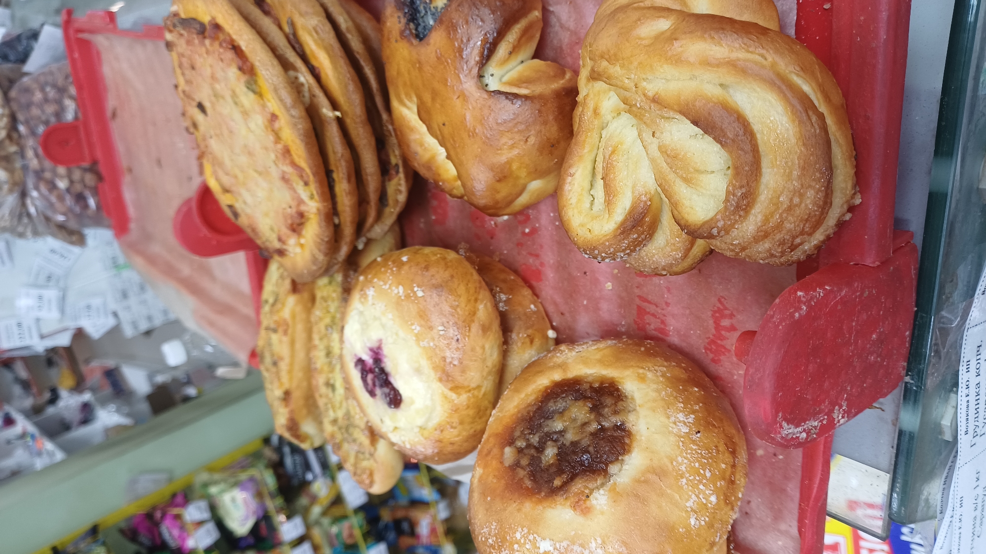 A close-up, slightly elevated, vertical shot shows a variety of baked goods displayed on a red paper liner and a red tray. The baked goods include several circular pastries topped with a creamy filling and berries, circular pastries with a dark, jam-like filling, and a stack of flat, savory pastries topped with cheese and what appear to be vegetables. There are also twisted, sweet buns dusted with sugar. In the background, shelves of products are visible, with some price tags. The scene appears to be inside a market or bakery in the city of Podgornoye, Russia. There are no people or interactions visible in the frame.