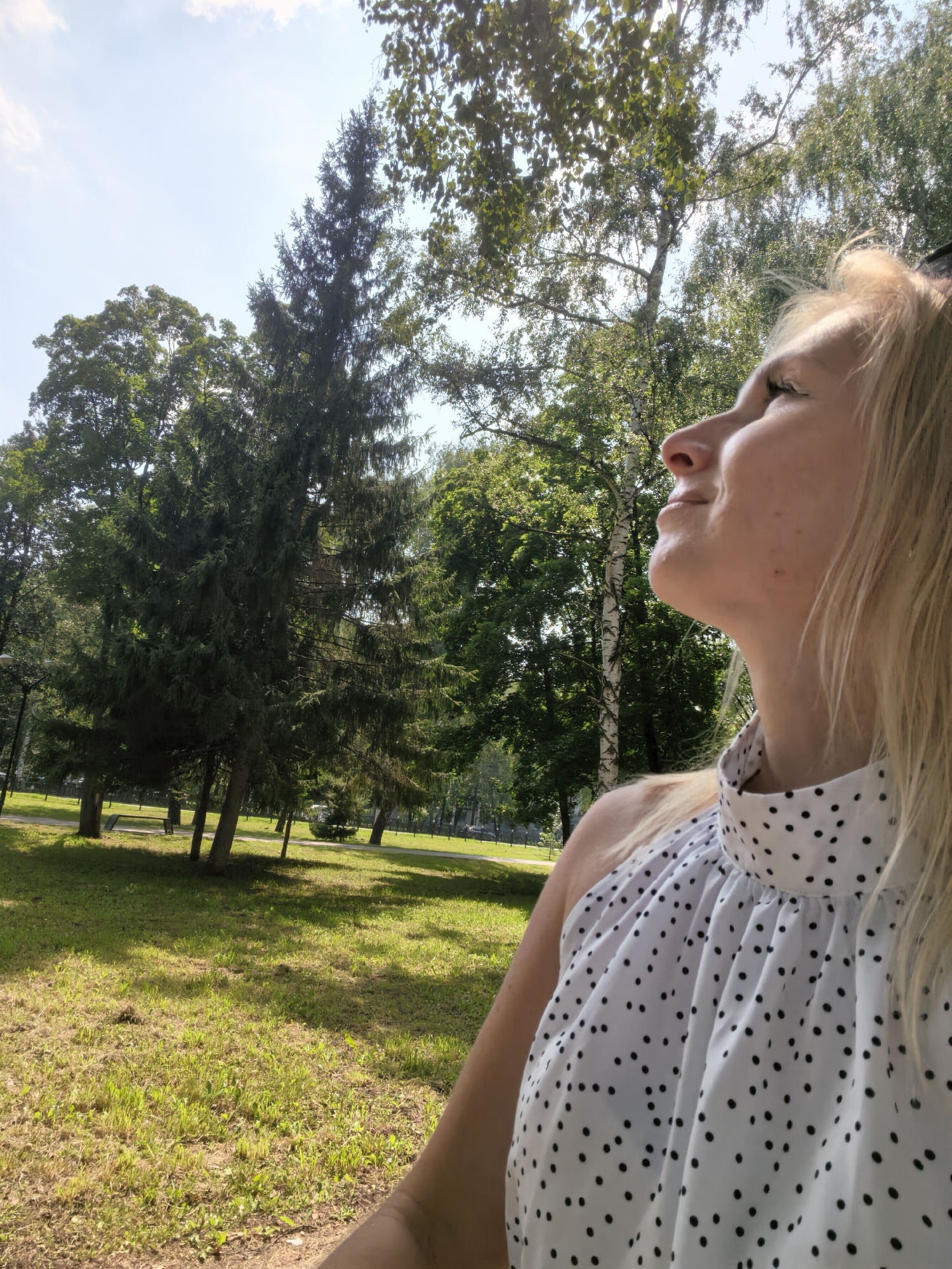A woman with blonde hair is shown in a profile view, looking upwards to the right of the frame. She is wearing a white halter top with a black polka dot pattern. The background consists of a park setting with lush green grass, various trees, and a clear blue sky with scattered clouds. The lighting suggests it is a sunny day. The specific location in the city of Sterlitamak, Russia, cannot be determined from the visual information provided.
