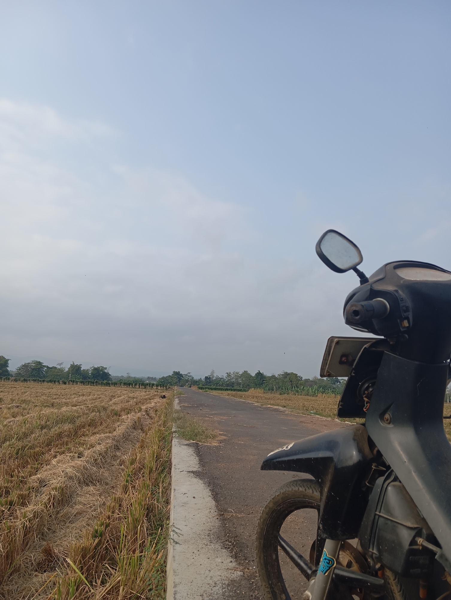 The image depicts a rural road in Sokaraja, Indonesia, with a portion of a black motorcycle in the foreground. The motorcycle, positioned on the right side of the frame, shows its front wheel, fender, and part of its body. The road is paved and has a concrete curb. To the left of the road, a field of dry, harvested crops, likely rice straw, stretches towards a line of trees in the middle distance. The sky is overcast with a pale blue hue and a few faint clouds. The overall scene suggests a dry season in a rural agricultural area. There are no people visible in the image.