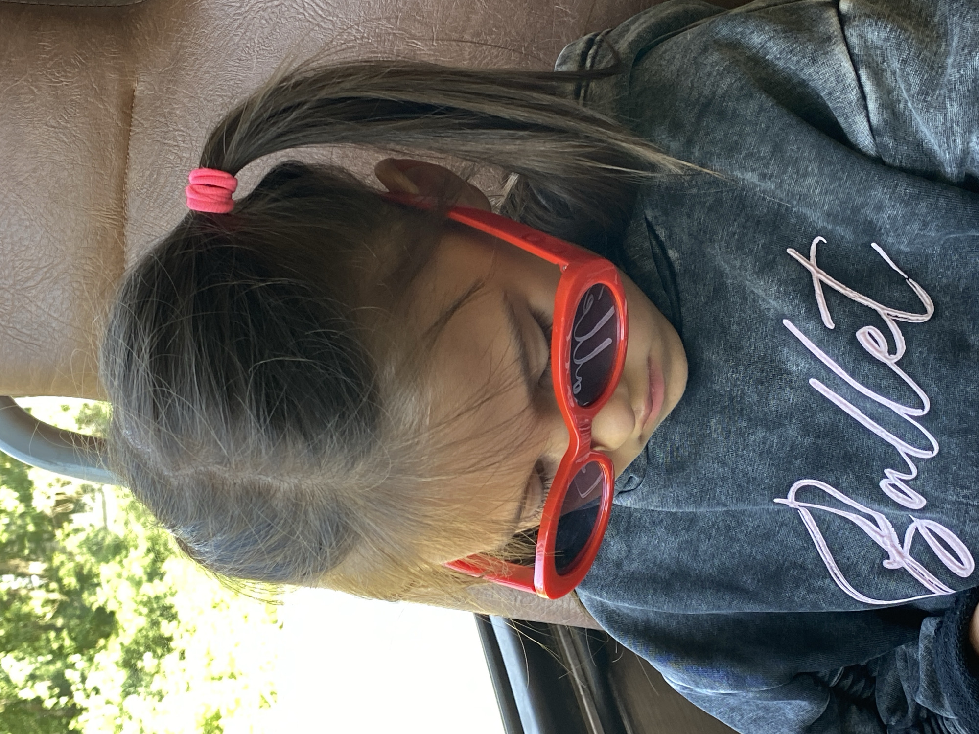 A close-up, waist-up, indoor shot shows a young girl wearing red oval-shaped sunglasses and a dark grey hoodie with "Ballet" written on it in pink cursive. Her hair is pulled back into two pigtails with pink hair ties. She is looking down, possibly at a device not visible in the frame. The background is out of focus, showing a blurry green outdoor scene and a brown textured surface, possibly a seat. The lighting suggests it's daytime. The location of Chaikovsky, Russia is not evident from the image.