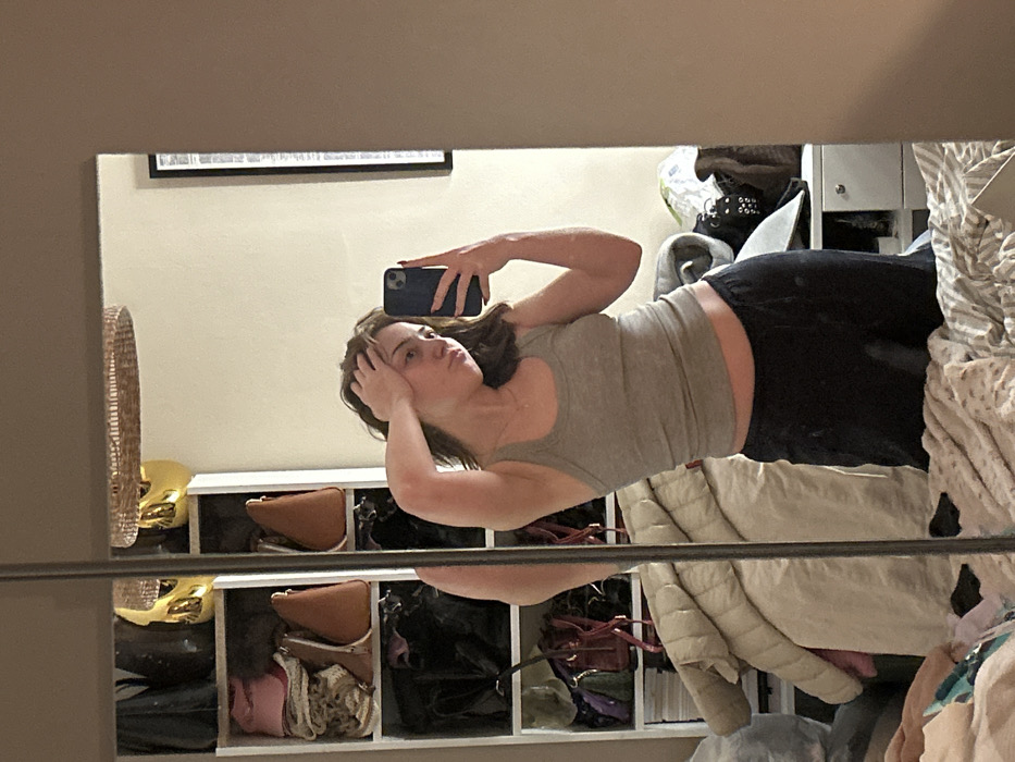 A young woman in a gray tank top and black leggings holds a phone to take a selfie in a mirror. She stands in front of a white shelving unit filled with bags. A framed picture hangs on the wall above the shelving. A wicker basket is visible to the left. The right side of the reflection shows clothing piled on a bed or surface.