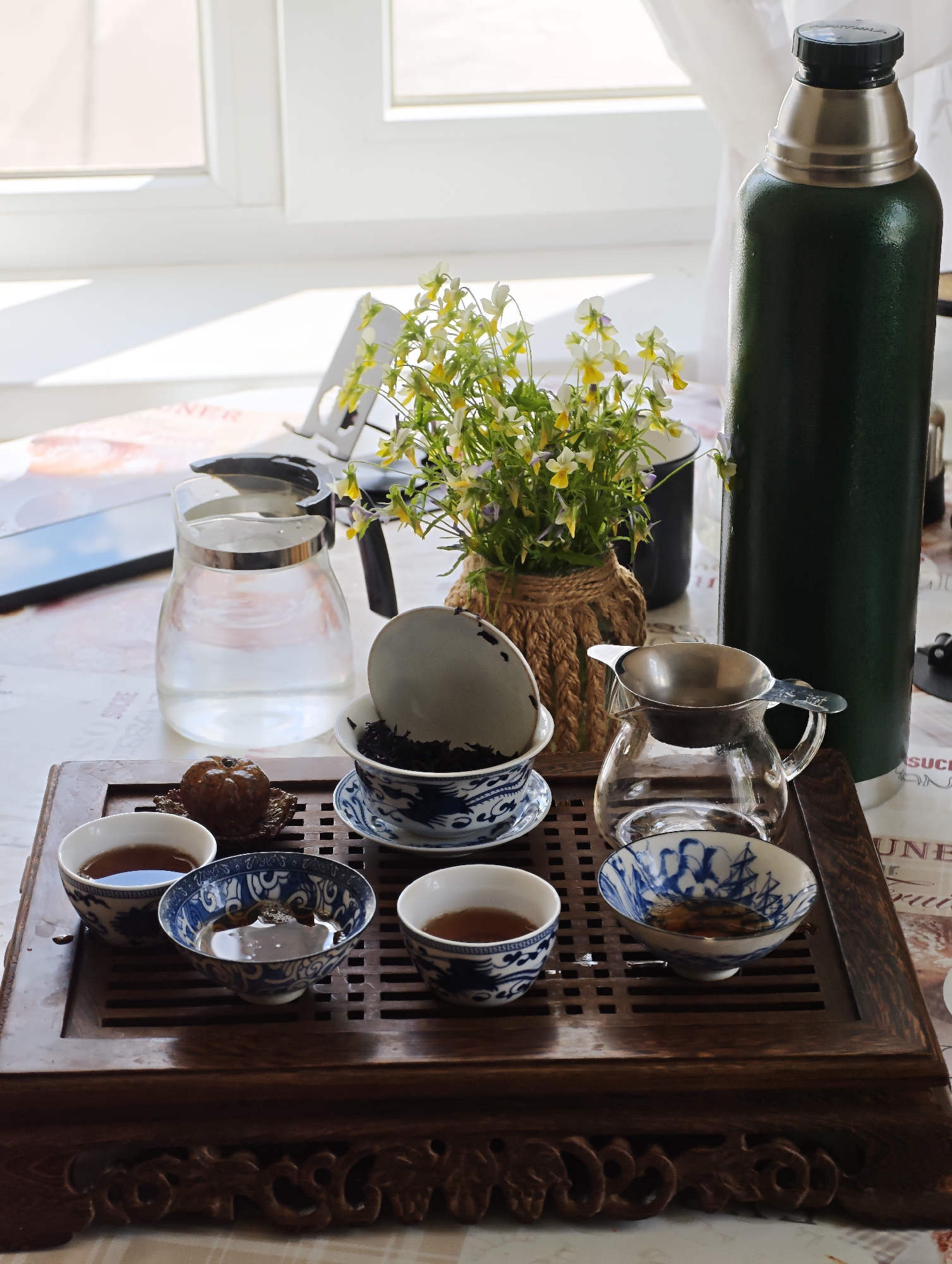The image depicts a traditional Chinese tea ceremony setup on a wooden tray placed on a table. The tray holds several items, including a glass pitcher filled with water, multiple small porcelain teacups with blue and white Chinese patterns, a teapot with a metal strainer, and a small decorative item resembling a dried fruit. A dark green thermos stands upright to the right of the setup. In the background, a bouquet of small yellow and white flowers in a woven pot is visible, along with a window and white curtains. The scene appears to be indoors, likely in a domestic setting. The presence of a thermos suggests that hot water is available for brewing tea. The overall atmosphere is serene and suggests a moment of quiet contemplation or a social gathering centered around tea. The image does not contain any people or animals, and there are no discernible actions or interactions taking place. The lighting suggests it is daytime, with light coming from the window. The location, Парэчча, Belarus, is provided but there is no visual information within the image to confirm this specific location.