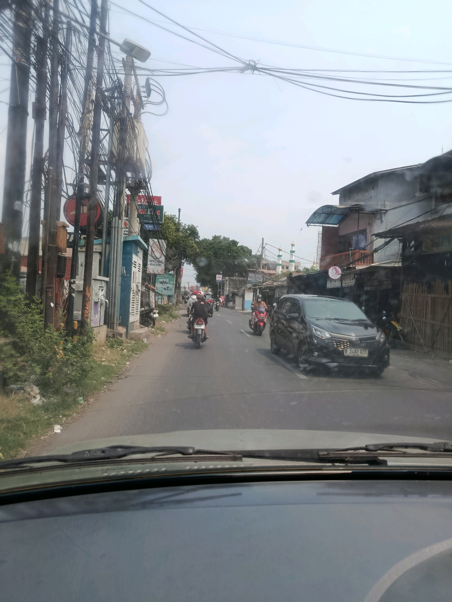 This image captures a street scene in East Jakarta, Indonesia, viewed from inside a vehicle. The road is lined with buildings and a dense network of utility poles and wires on the left side. Several motorcycles and a black car are on the road. The car, a Toyota Avanza, has the license plate B 1483 DZO. On the right side of the road, there are shops and houses, with bamboo fencing visible. In the background, a mosque with minarets can be seen. The sky is overcast. The foreground shows the dashboard and windshield of the vehicle from which the photo was taken.