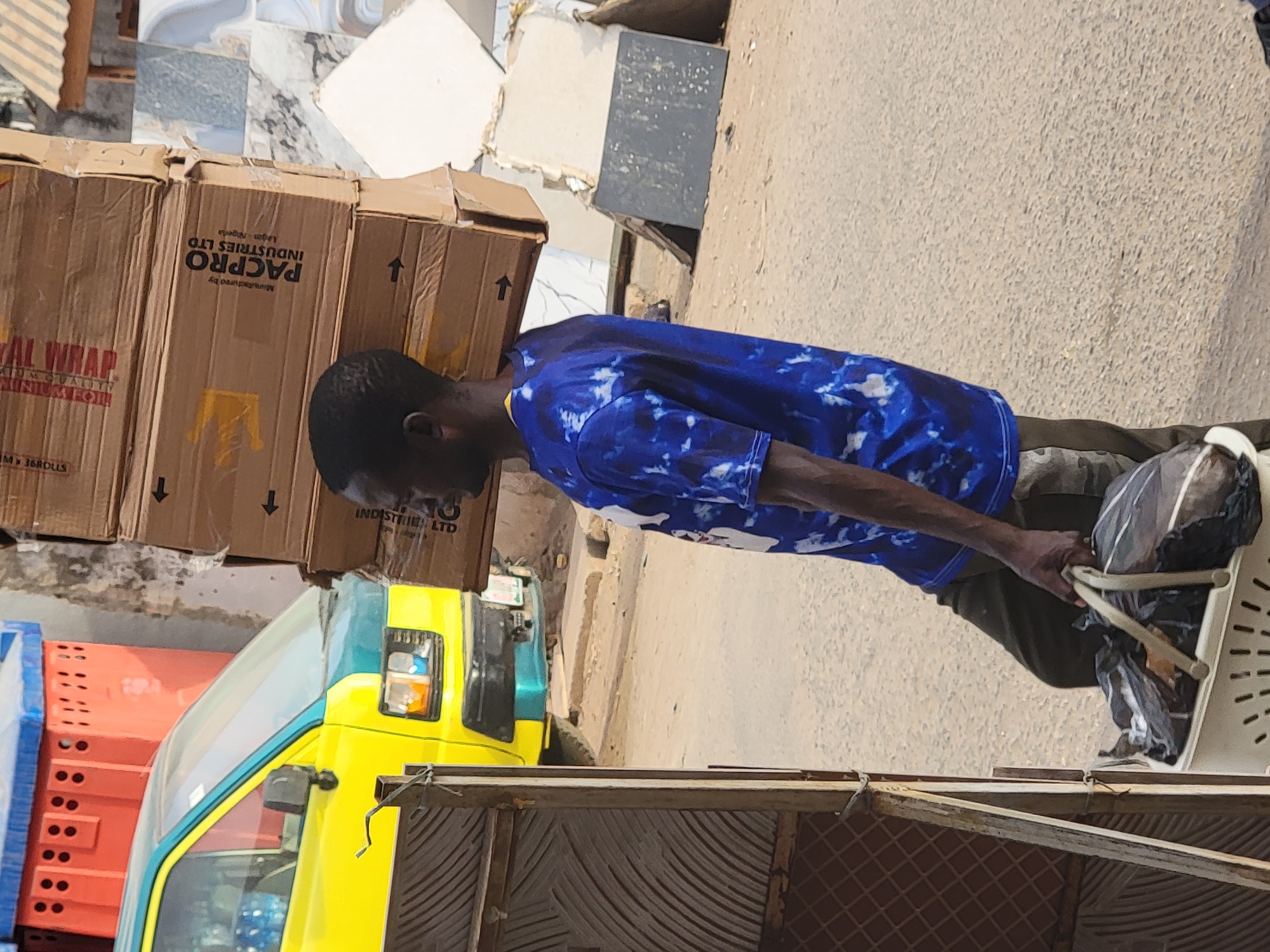 A man is carrying a load of cardboard boxes on his head. The boxes have the words "PACPRO INDUSTRIES LTD" and "AL WRAP" printed on them. A yellow truck with a blue trim is visible in the background, next to a stack of orange plastic crates. The scene appears to be in an urban environment, likely Maiburiji, Nigeria, based on the printing on the boxes. The photo appears to be taken from a slightly elevated vantage point, possibly from behind a fence.