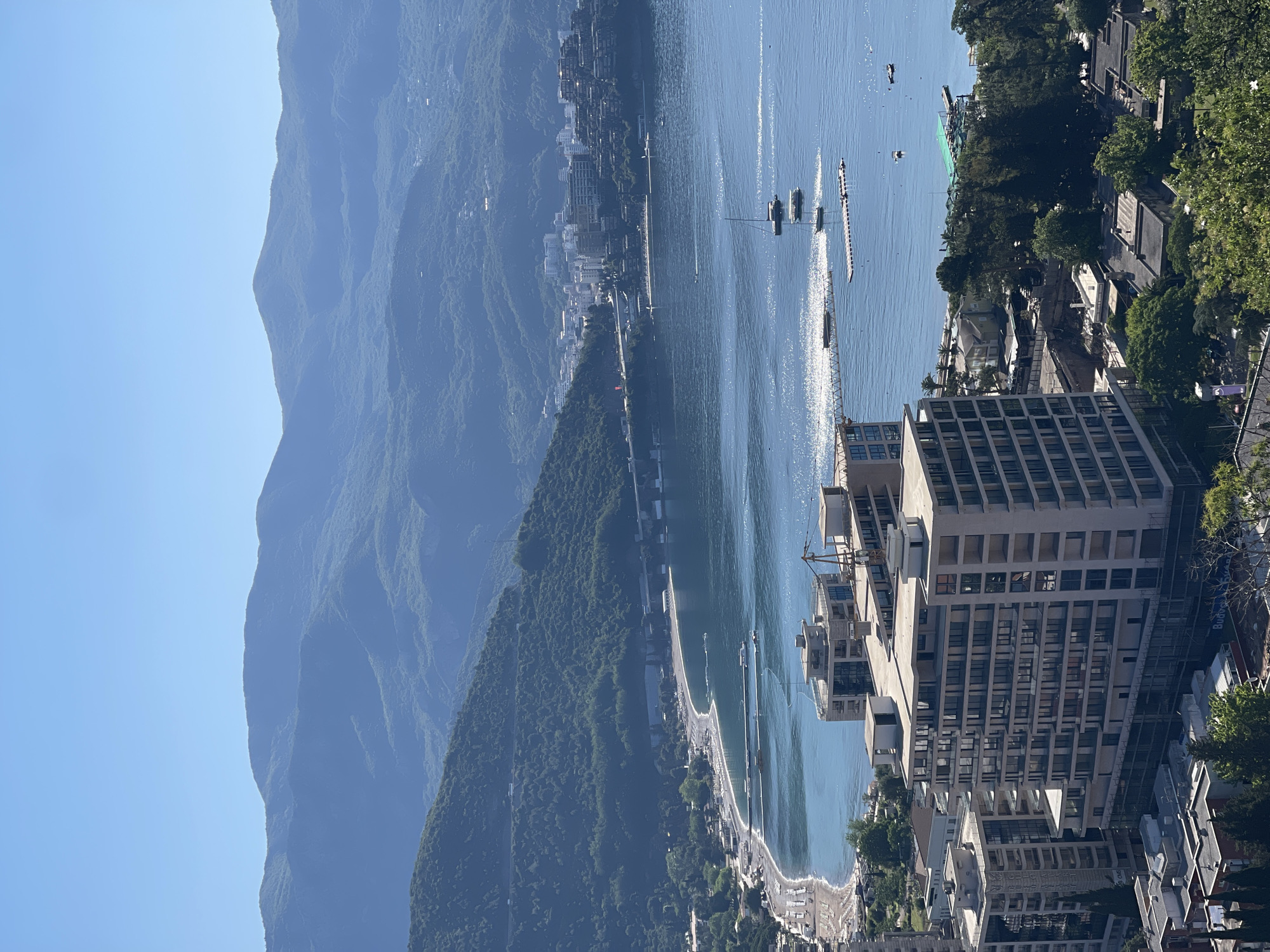 The image shows an elevated, slightly oblique view of Budva, Montenegro, with mountains in the background, a large body of water (likely the Adriatic Sea), and a cityscape in the foreground. A prominent modern building with many windows dominates the lower right of the frame. A long stretch of beach and a pedestrian walkway are visible along the shoreline. There are several boats and smaller vessels on the water, some of which are creating wakes. The mountains are forested and hazy. The sky is clear blue.