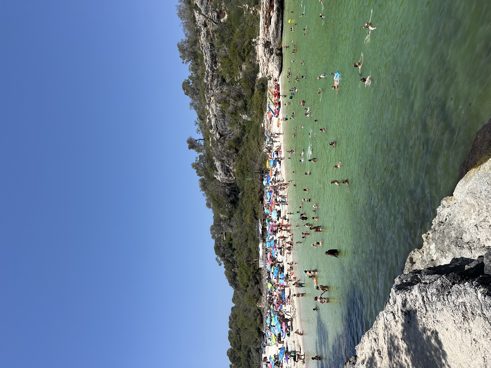 This is an eye-level, full shot of a crowded beach scene on a sunny day. The image is oriented vertically, with the sky on the left and the beach and water on the right.
The beach itself is sandy and lined with numerous colorful umbrellas and beach chairs, indicating a leisure activity is taking place. Many people are sunbathing, sitting, or walking on the sand. The water is a clear, greenish hue and is populated by a large number of swimmers. Some are wading near the shore, while others are swimming further out. A few individuals are visible on inflatable tubes.
Behind the beach, a steep, rocky cliff covered in dense green foliage rises up. The cliff face is rugged and uneven, with trees growing out of its crevices. The overall impression is a popular tourist destination during peak season. The clear blue sky and bright sunlight suggest it is midday, with pleasant weather. There are no visible signs or text in the image.