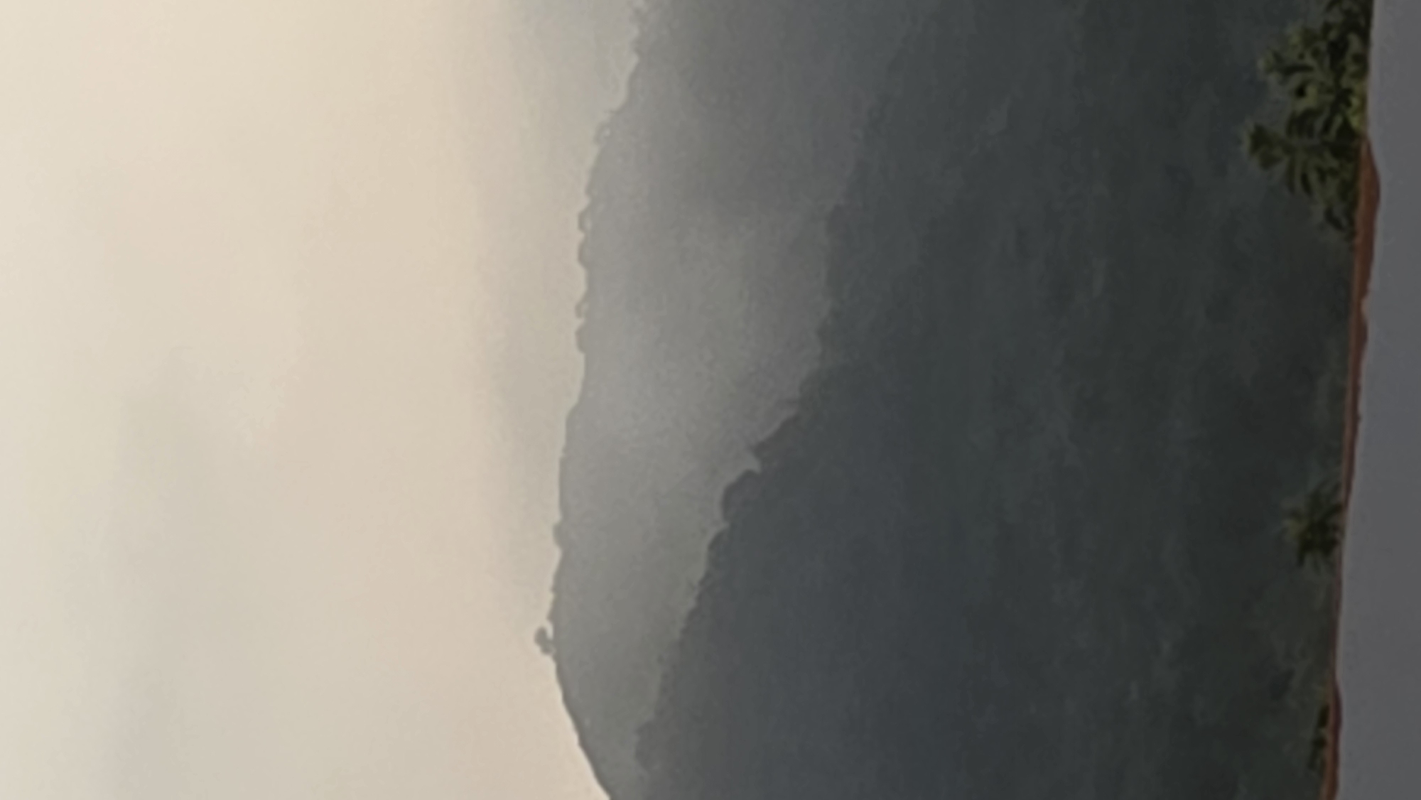 The image presents a hazy mountain landscape, possibly in the Bac Natural Heritage Area Trang, Vietnam, with the mountains appearing in shades of gray and brown due to fog and distance. The right side of the image features trees, while the left side is obscured by fog. The entire scene exudes a quiet, hazy atmosphere.