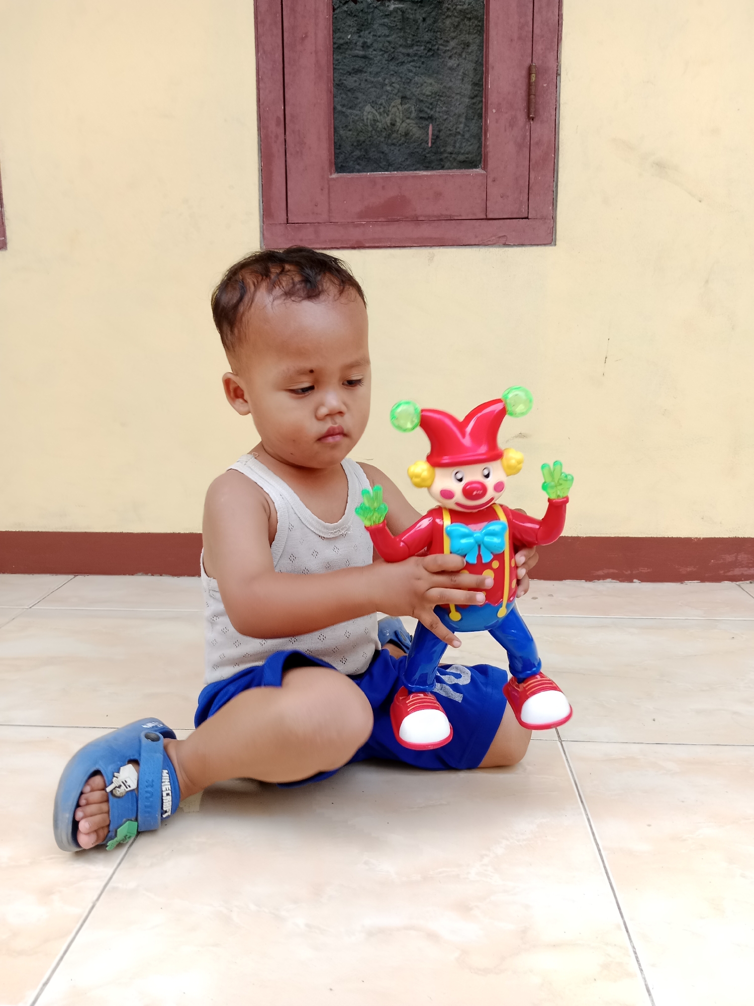 A young child, possibly a toddler, sits on a tiled floor in front of a light yellow wall with a dark brown baseboard and a wooden-framed window. The child wears a white tank top and blue shorts, with blue sandals that appear to have a Minecraft theme. They are holding a red and blue toy clown with a blue bow tie and yellow accents. The child's left hand is on the clown's body, and their right hand is near the clown's head. The background wall is plain, with some smudges and discolorations. The window has a dark, textured glass. The lighting suggests it is daytime. The image appears to have been taken outdoors, possibly in a courtyard or patio area. The location is Parage, Indonesia.