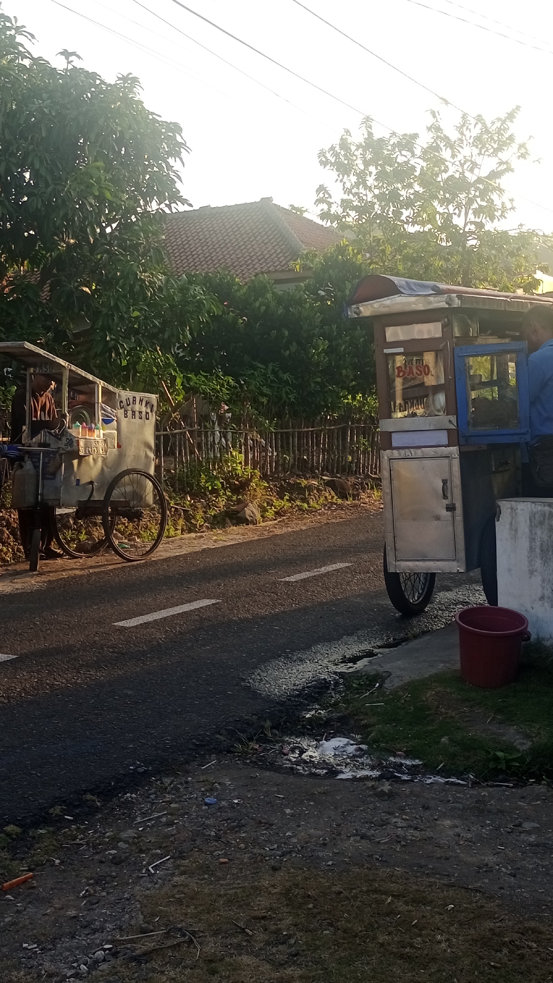 The image depicts an outdoor scene with two food carts parked on the side of a paved road. The food carts appear to be mobile, one with two large wheels and another with a single large wheel. The cart on the left has text that appears to read "CUANKI BASO" on its side. The cart on the right has the word "BASO" prominently displayed. Trees and a house with a tiled roof are visible in the background. The lighting suggests it is either early morning or late afternoon. A red bucket is visible near the food cart on the right, and some litter is present on the ground near the road. The scene suggests a street-side food vendor operation in a residential or semi-rural area, possibly in Pakemitan, Indonesia.