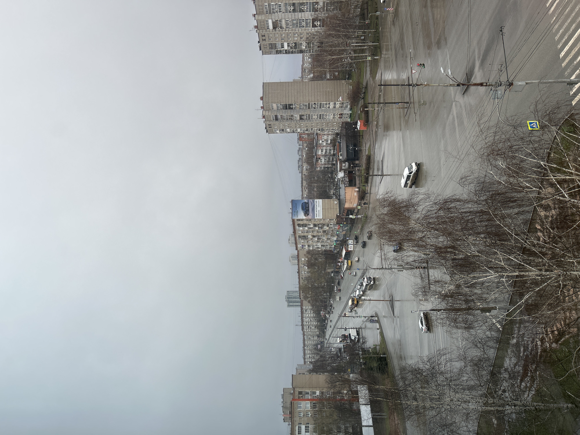 An aerial view captures a wide urban street in Izhevsk, Russia, on an overcast and wet day, likely late autumn, winter, or early spring. The sky is a uniform dull grey, reflecting off the damp, glistening asphalt of the multi-lane road.
The street, appearing to be a main avenue, is populated with moving and parked cars, suggesting normal daily traffic. A notable white car is parked or temporarily stopped partially in a shallow puddle or standing water at the roadside in the foreground. Bare deciduous trees with intricate, leafless branches line the sides of the road and fill green spaces, indicating the colder season.
On both sides of the street, multiple multi-story apartment buildings with numerous windows, characteristic of Soviet-era architecture, stretch into the distance. Some buildings show signs of wear or open windows. Utility poles, power lines, streetlights, and traffic signals are scattered along the thoroughfare. Crosswalk markings are visible on the wet road surface.
Visible text on a prominent billboard on the right side of the street (from the viewer's perspective) advertises a car dealership. The Cyrillic text reads "ЦЕНТР КОММЕРЧЕСКОГО ТРАНСПОРТА" (Center of Commercial Transport) and mentions "Официальный дилер ГАЗ" (Official GAZ dealer), with a website "цкт.рф". Other road signs include a yellow pedestrian crossing sign. The overall impression is one of a bustling city scene under grey, cool, and damp weather conditions.