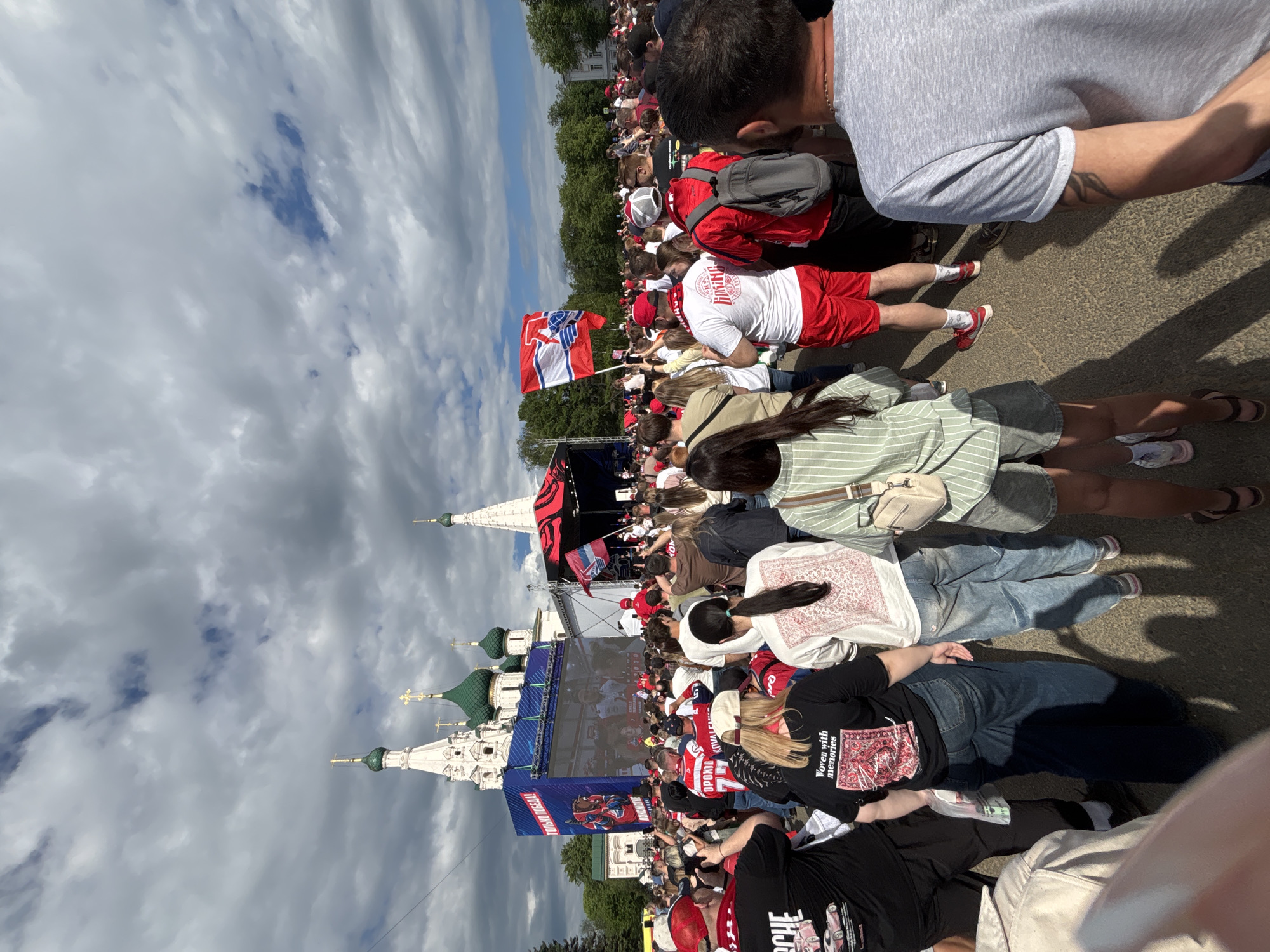 A crowd of people is gathered outdoors in Rusanovo, Russia, facing a large screen and stage with the image of an ongoing sport event and flags of the national ice hockey team. The Church of Elijah the Prophet stands on the left side of the frame, under a sky with clouds. The crowd is comprised of people of various ages. Some are wearing red and white clothing. Some people are sitting, while others are standing. The ground is paved.