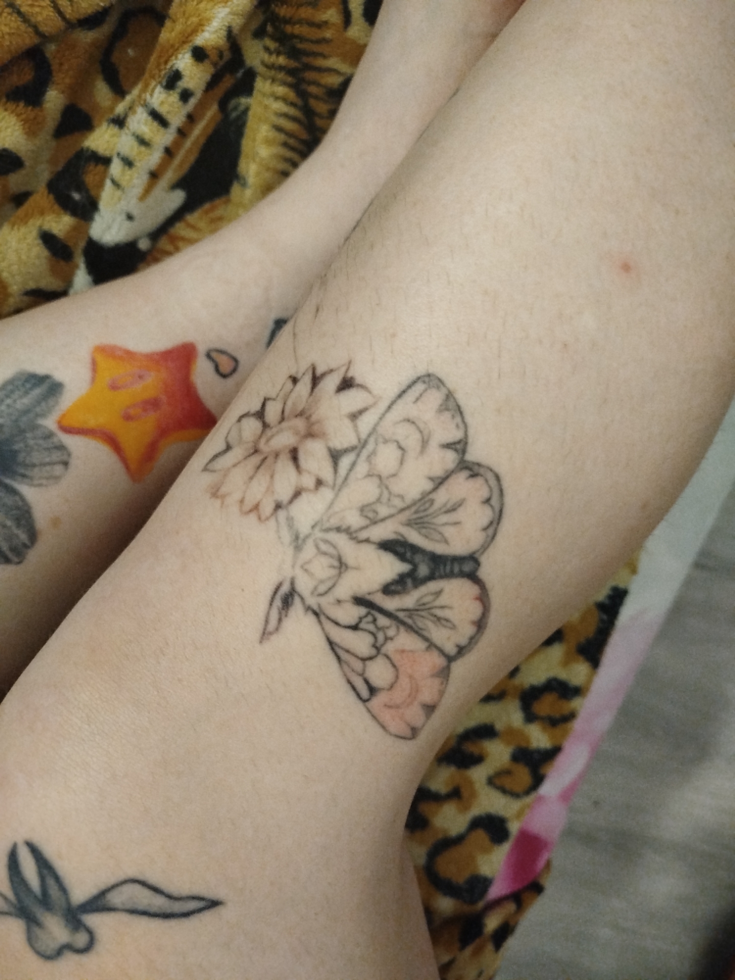 The image shows a close-up of a person's leg. Several tattoos are visible, including a flower with a moth on the thigh and a star on the leg in the background. The leg rests against a patterned cloth. The location is Unknown, Unknown.