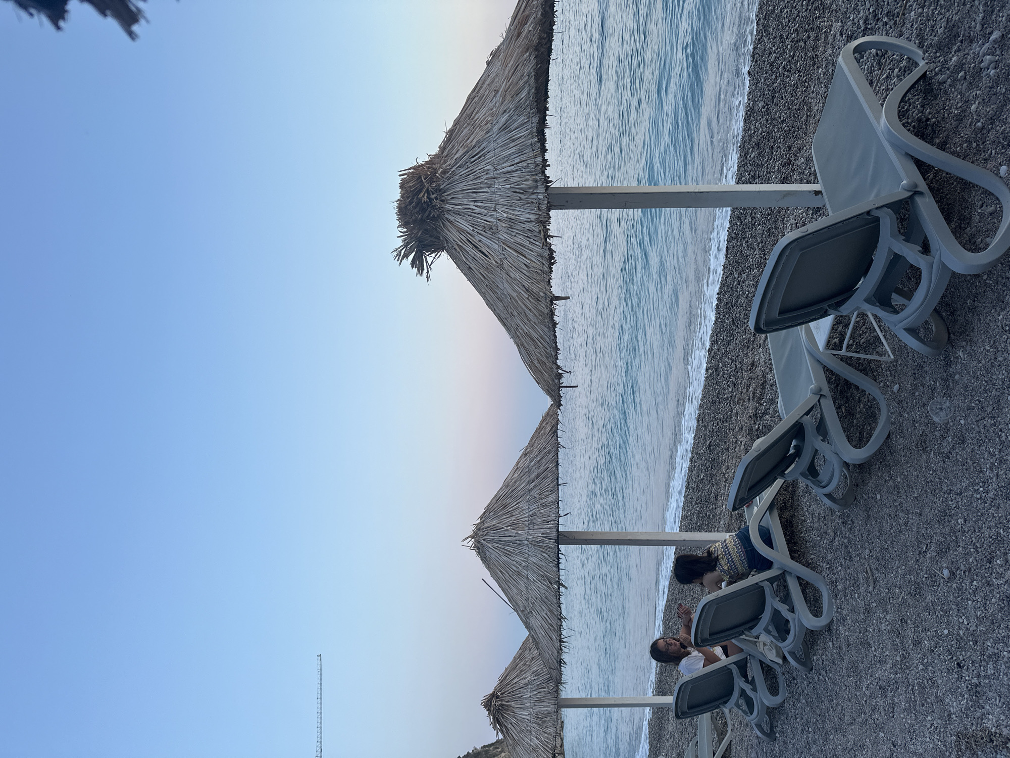The image depicts a serene beach scene in Mirište, Montenegro, at dusk. In the foreground, several grey plastic lounge chairs are arranged on a pebble beach, some unoccupied and others occupied by two individuals. The first person, facing left, is wearing a white t-shirt and dark-rimmed glasses. The second person, facing right, is wearing a patterned top and dark pants. They appear to be engaged in conversation. Behind the lounge chairs are three thatched umbrellas, providing shade. The calm, blue sea with gentle waves is visible in the background, stretching towards the horizon under a clear, pale blue sky that transitions to soft pink hues near the horizon. A faint, distant crane is visible in the upper left corner of the sky. The overall atmosphere is tranquil and relaxing.