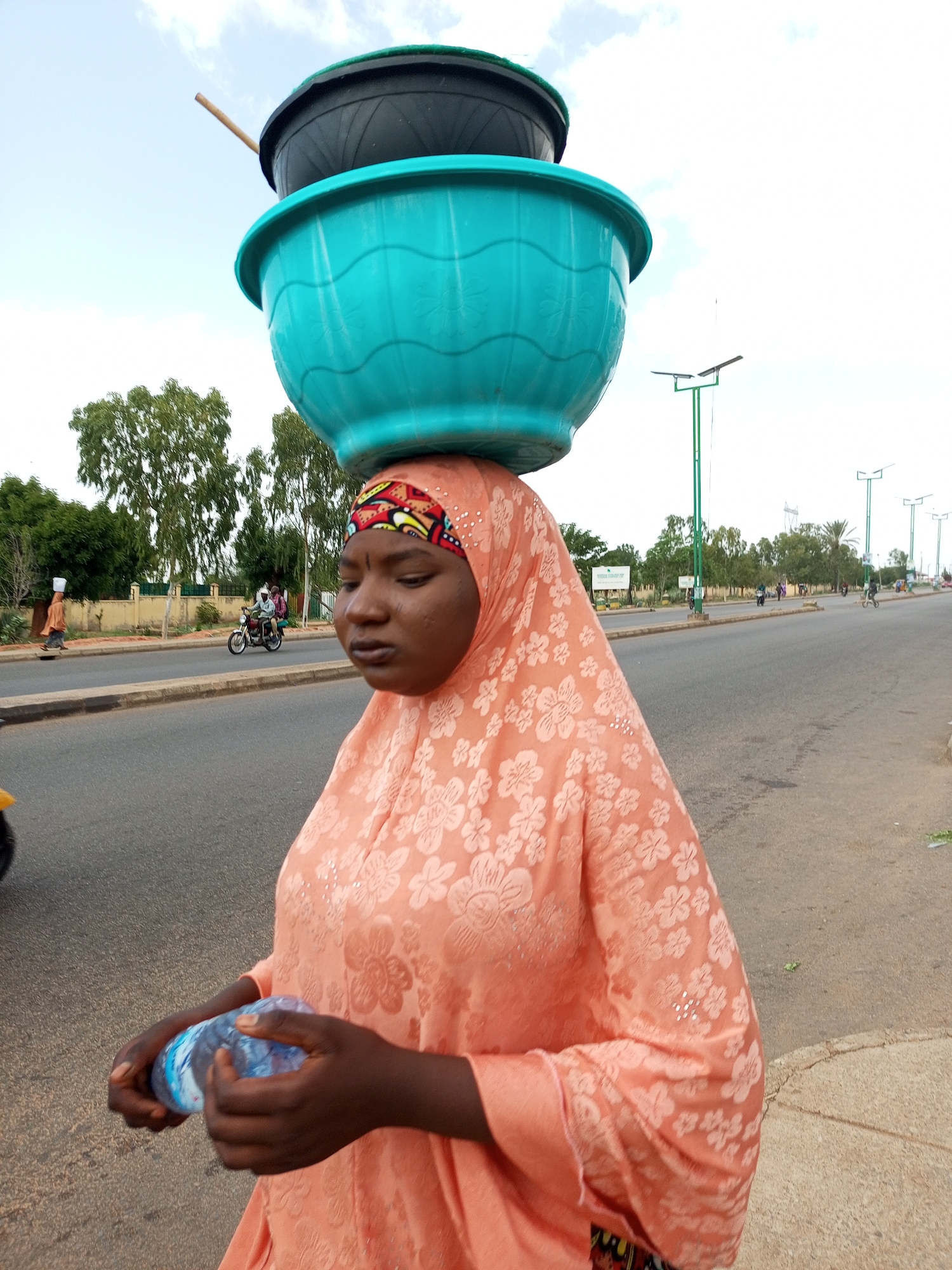 A woman in Gombe, Nigeria, walks along a street with two large plastic basins stacked on her head. She is wearing a peach-colored headscarf and a peach-colored dress with a floral pattern. In her hands, she holds a plastic bottle of water. The street is paved and appears to be a main thoroughfare, with a sidewalk on the right side of the image. Trees line the background, and there are several streetlights visible. In the distance, a motorcycle with two passengers is seen on the road, and another person is walking on the sidewalk in the far background. The sky is overcast with some clouds.