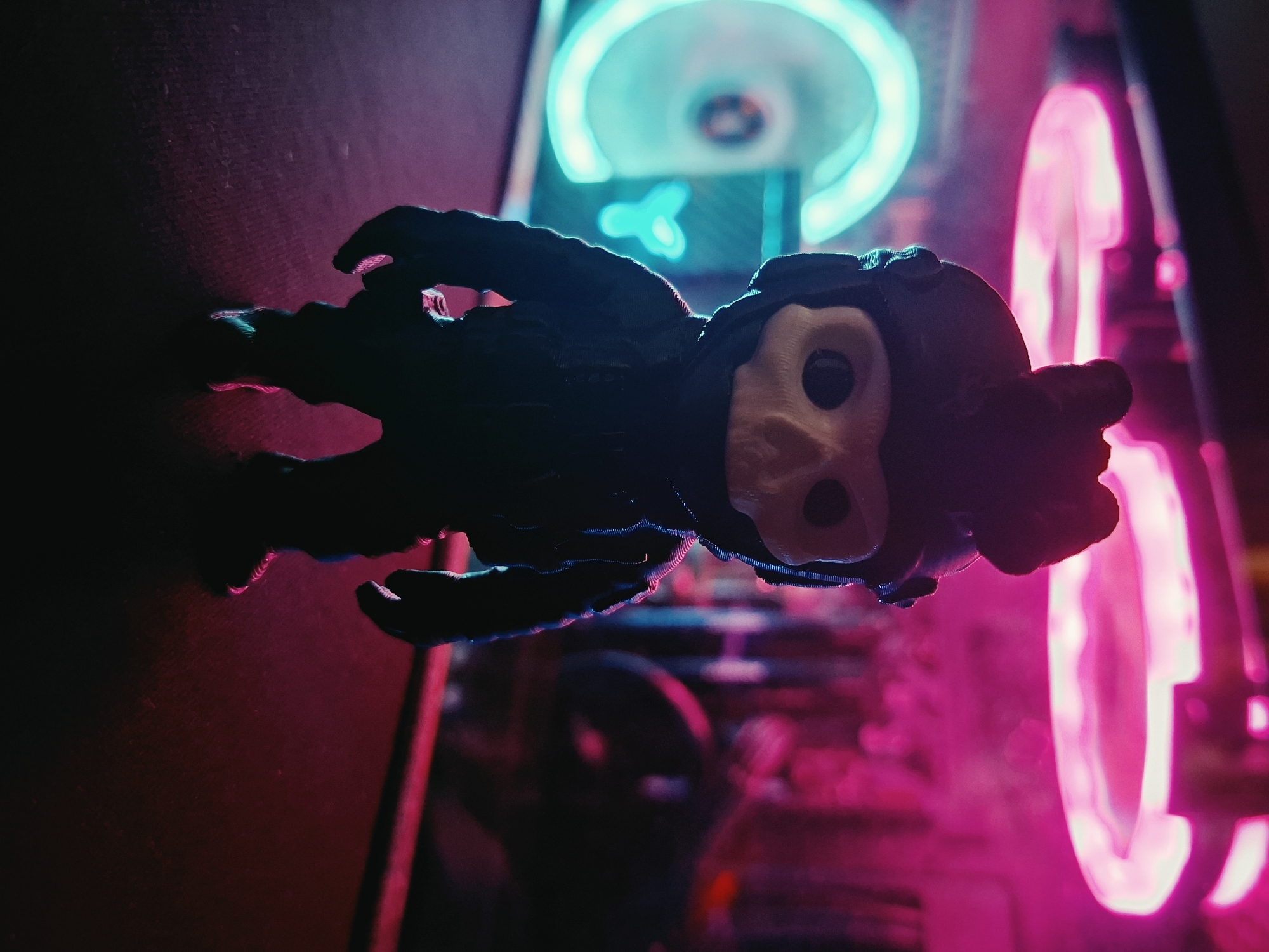 A dark, low-angle, close-up shot captures a stylized, skull-faced figurine with a tactical helmet and suit. The figurine stands on a dark, textured surface. In the background, a computer case is visible, illuminated by vibrant neon pink and cyan lights. The pink lights are from fans at the top of the case, and the cyan light is from a fan on the left side, which has a visible "CY" logo. The scene is dimly lit, with the neon lights casting colorful reflections on the figurine and the surrounding environment. The overall atmosphere is moody and futuristic. The image does not contain any information about the city of Vladivostok, Russia.