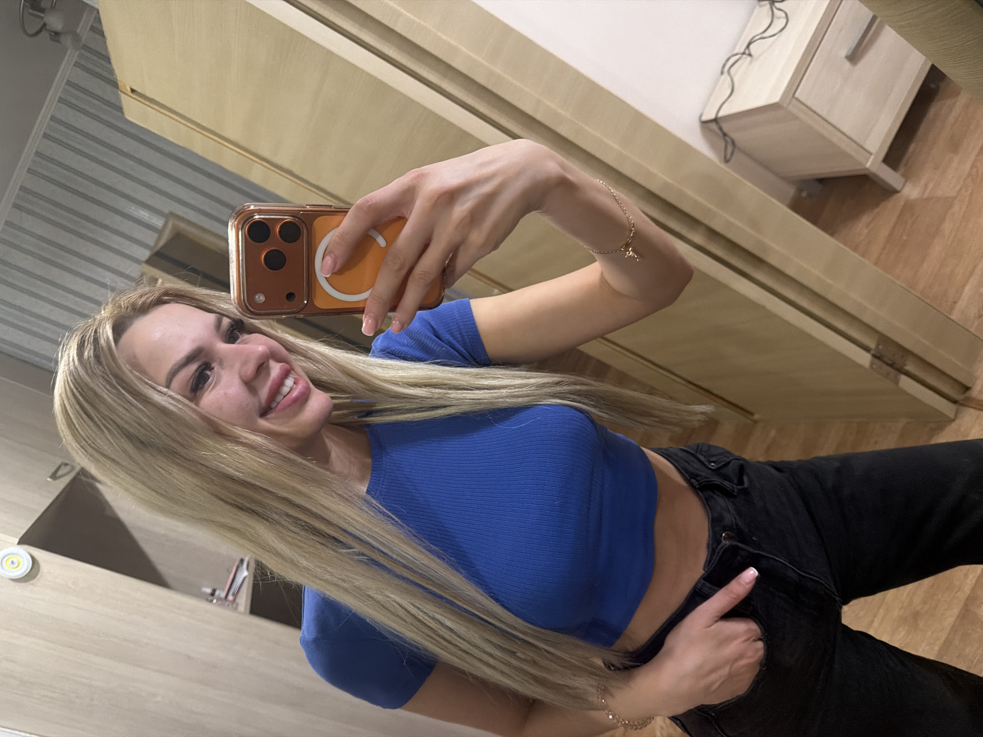 A young woman with blonde hair is taking a mirror selfie in an apartment. She is wearing a blue crop top and black jeans, and a bracelet on her left wrist. She is smiling and holding a smartphone in her right hand. The background shows a wardrobe, a nightstand with a light cord, and a wooden floor. The image does not provide any specific information about the location in Kyiv, Ukraine.