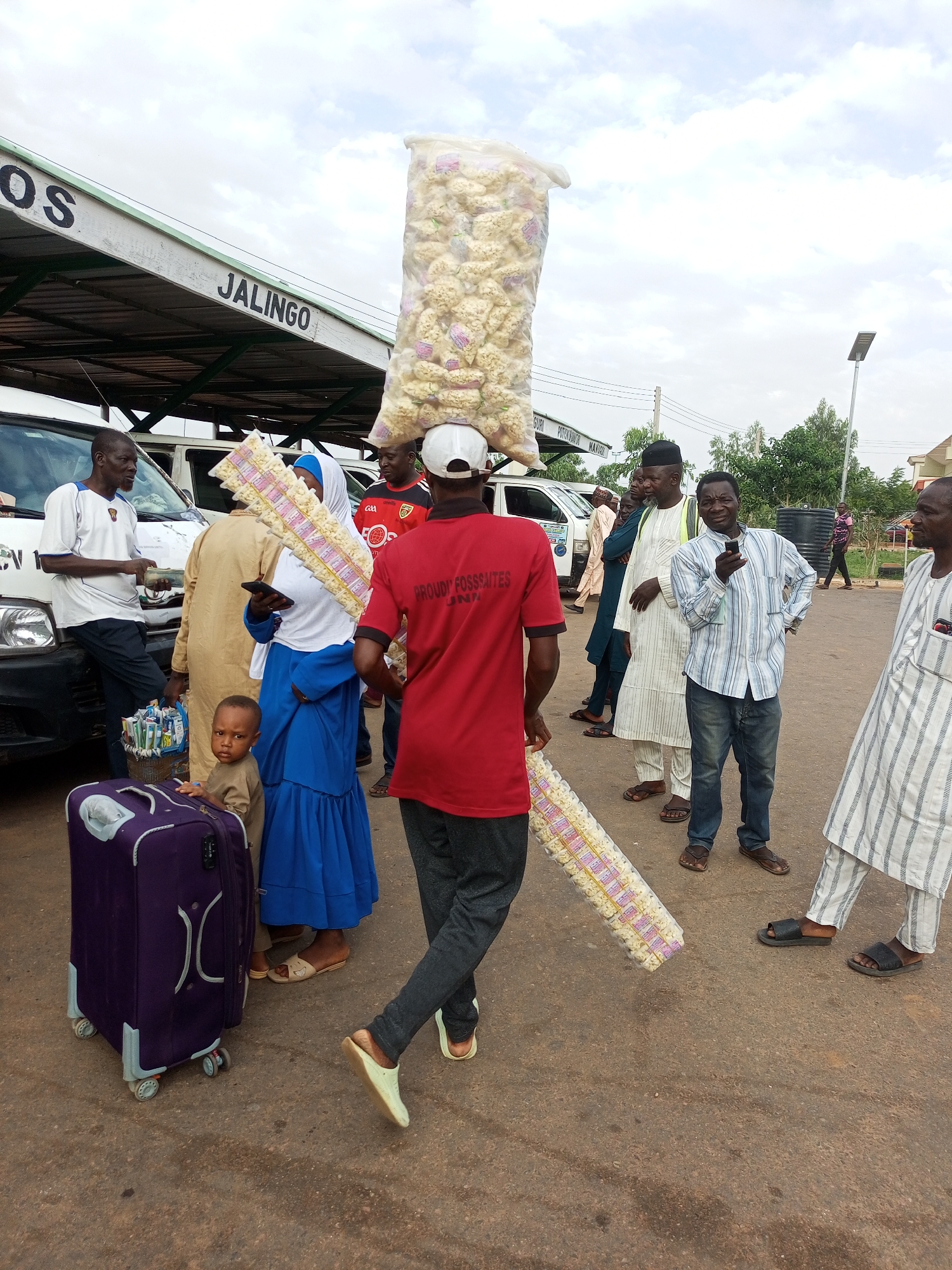 In the city of Gombe, Nigeria, a group of people are gathered outdoors, likely at a transport terminal. A man in a red shirt with white lettering that reads "PROUDLY FOSSAITES" is carrying a large, clear plastic bag filled with small, light-colored items on his head. He also holds a long, flat item wrapped in plastic, which appears to be several packages of snacks or food items. To his left, a woman in a blue dress and hijab is holding a mobile phone. Beside her, a young boy in a brown shirt stands next to a large, purple rolling suitcase. Other individuals are visible in the background, some wearing traditional Nigerian attire. A white bus with "JALINGO" written on its side is parked behind some of the people. The ground is paved with asphalt, and the sky is overcast. The scene suggests a moment of transit or commerce.