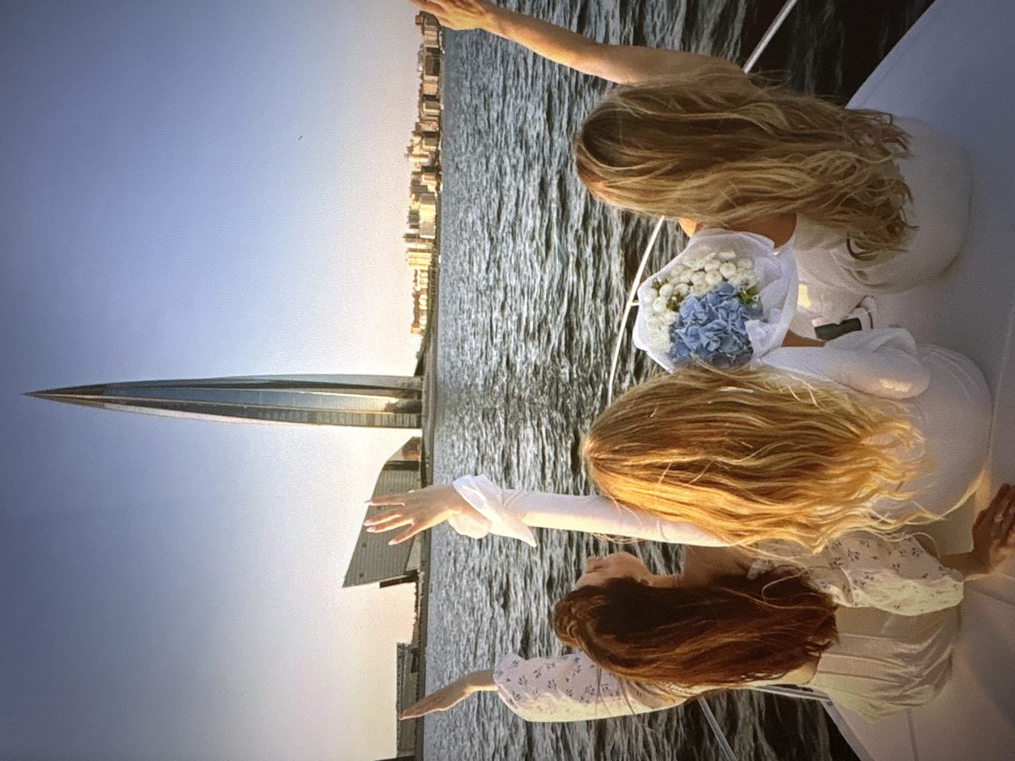 Three women are on a boat in the water, with a city skyline and a tall, modern skyscraper in the background. The woman on the left has long, reddish-brown hair and is wearing a white shirt with a floral pattern. Her left arm is raised, and her hand is open. The woman in the middle has long, blonde hair and is wearing a white top. Her arms are raised, and her hands are open. She is holding a bouquet of white and blue flowers. The woman on the right has long, blonde hair and is wearing a white top. Her right arm is raised, and her hand is open. The water is choppy, and the sky is clear and blue. The city appears to be St. Petersburg, Russia, due to the presence of the Lakhta Center skyscraper.