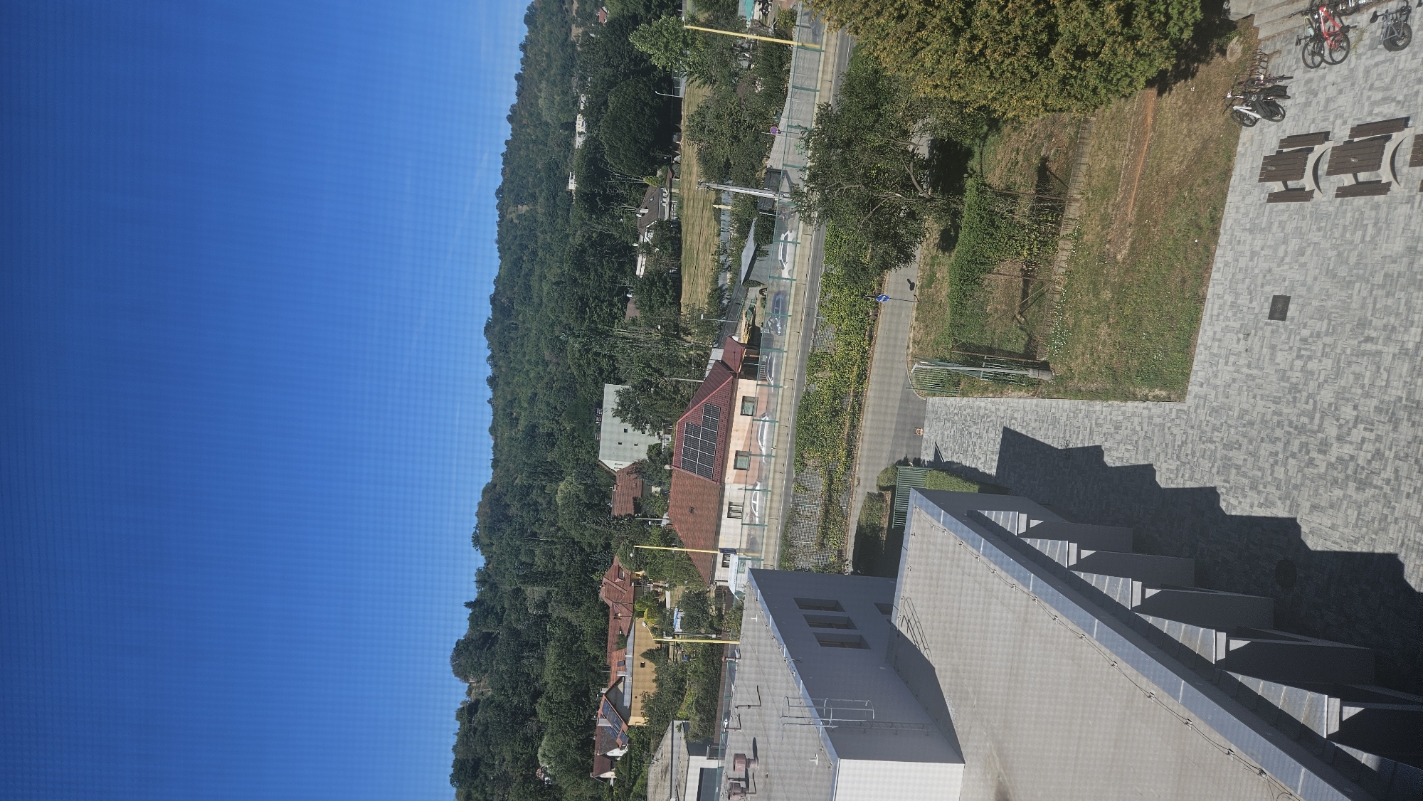 The image captures an elevated view of a residential area and a surrounding natural landscape, likely taken from a building. In the foreground, the roof and side of a modern building with a light-colored facade are visible. Adjacent to this building, a paved walkway leads past outdoor seating areas with wooden tables and benches, and a bicycle rack with several bicycles. The ground is covered with gray interlocking pavers, and a patch of grass with a few small white flowers is visible to the right.
Further into the scene, a road runs horizontally, with a protective barrier along its edge. Several cars are parked or driving on the road. Across the road, a house with a red tiled roof and solar panels is situated amidst greenery. Behind this house, a cluster of residential buildings with red and yellow roofs are nestled among trees.
The background is dominated by a dense, green, tree-covered hillside, suggesting the area is located on the outskirts of a city or in a valley. The sky above is a clear, bright blue. The overall scene depicts a tranquil, sunny day with a blend of urban development and natural scenery.
Based on the distinctive architectural style of the houses, particularly the one with the solar panels, and the characteristic topography of hills covered in dense forests, this scene is highly suggestive of the Zahorské vicinity in Prešov, Slovakia. This area is known for its suburban residential developments situated at the foothills of the Slanské vrchy mountains, offering views of both built-up areas and extensive greenery.