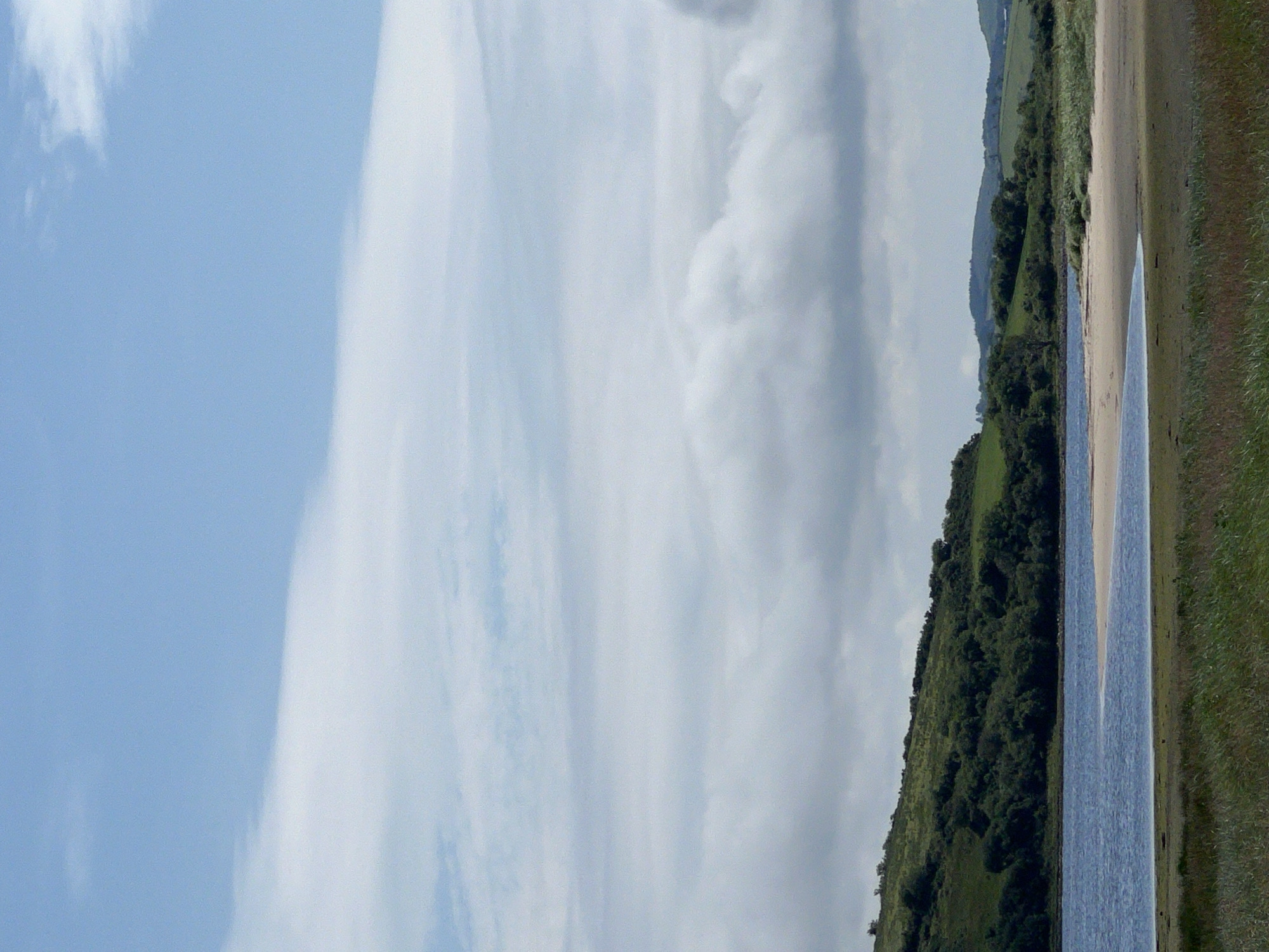 The image depicts a serene natural landscape, likely near Mountcharles, Ireland, under a partly cloudy sky. In the foreground, tall green grass lines a shallow body of water. A sandy beach curves into the water on the right side of the frame. Beyond the water, a lush green hillside is covered in dense trees and vegetation. In the distance, rolling hills form the horizon, with more scattered trees and green fields. The sky is a pale blue, with streaks of white clouds stretching across the upper portion of the image, some of which are quite dense and voluminous. There are no people or man-made structures visible in the scene, emphasizing the untouched, natural beauty of the environment.
