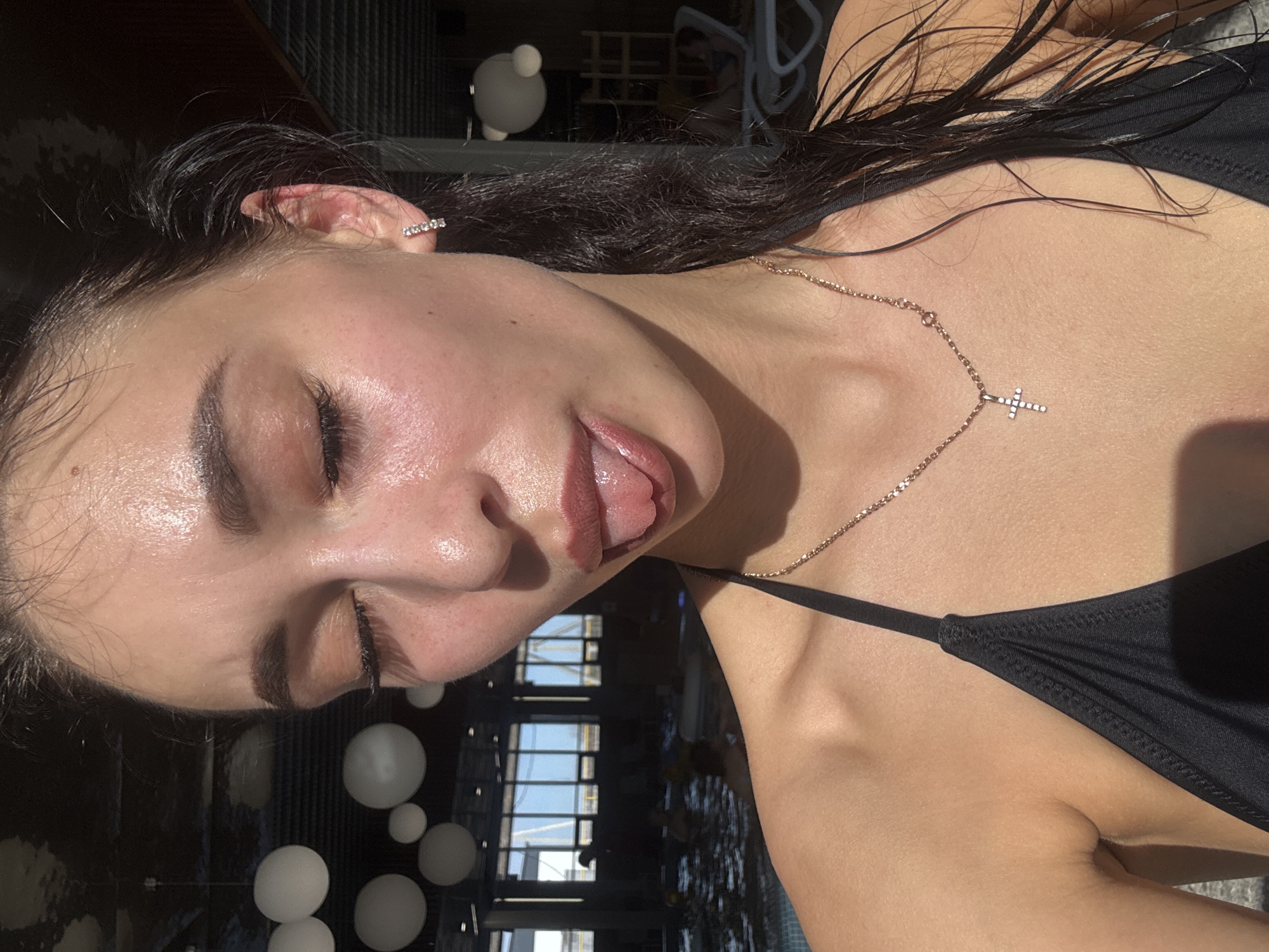 A woman is visible in a portrait shot. She has dark hair that appears wet, and her eyes are closed. Her tongue is sticking out slightly. She is wearing a black top with thin straps and a gold necklace with a small cross pendant. An earring is visible on her right ear. In the background, there is an interior with a window or opening showing a sky, and some spherical light fixtures are visible. Due to the lack of identifiable information in the image, determining the exact location in Votkinsk, Russia, is impossible.